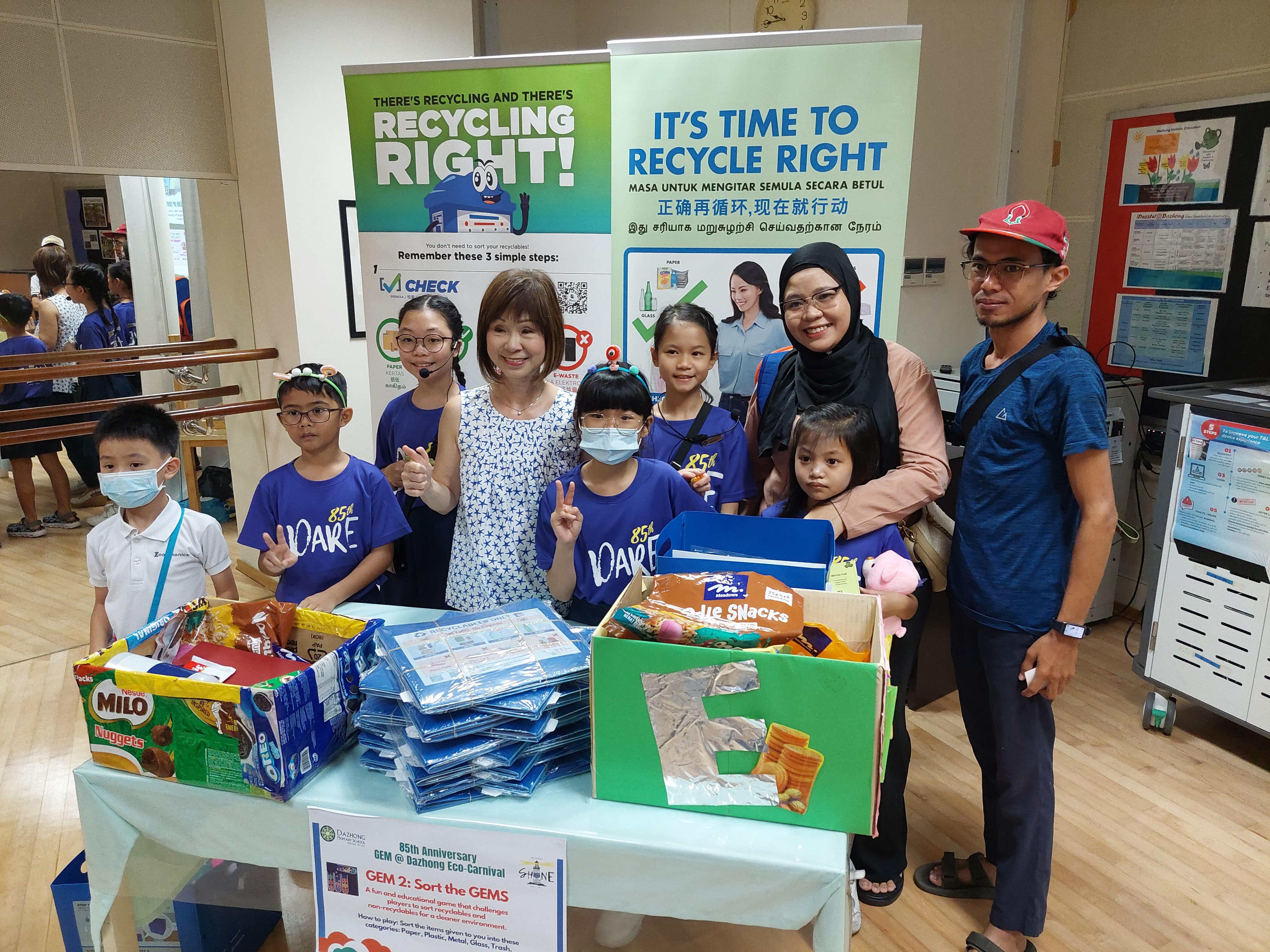 A group of adults and children pose at a recycling awareness booth with banners saying "Recycle Right."