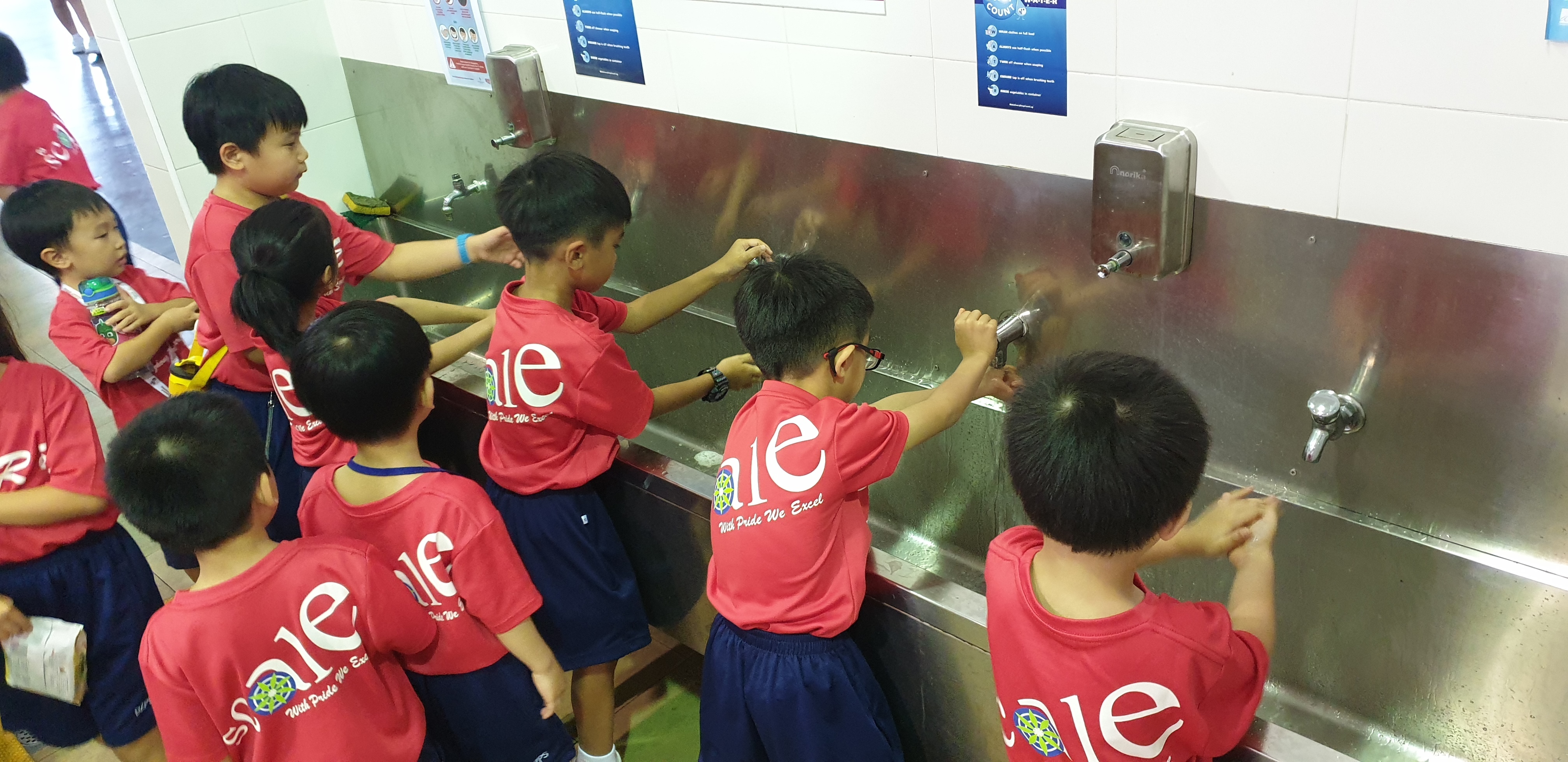 Young children in matching red school shirts crowd around a stainless steel trough sink, washing hands.