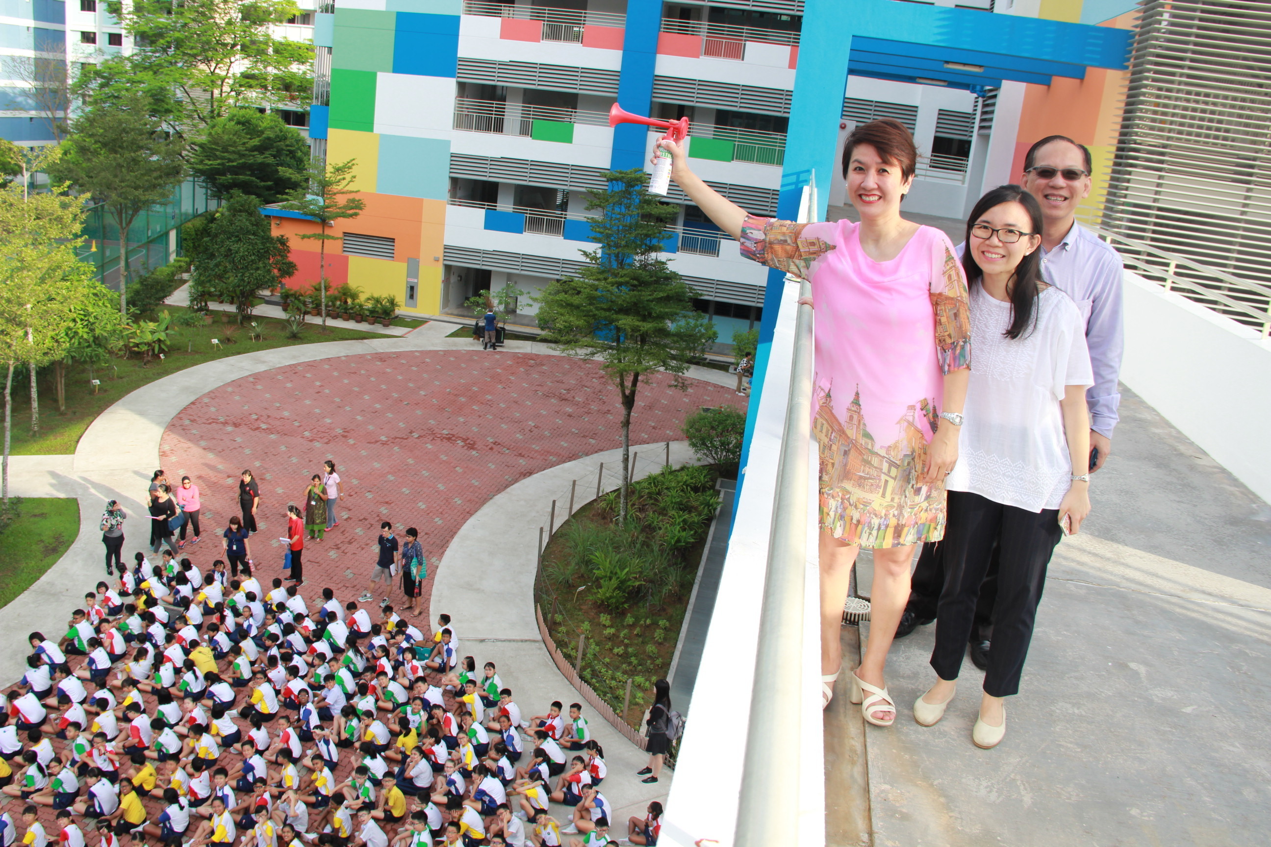 Three adults stand on a balcony above a schoolyard full of seated children in uniforms, one adult holding an air horn.