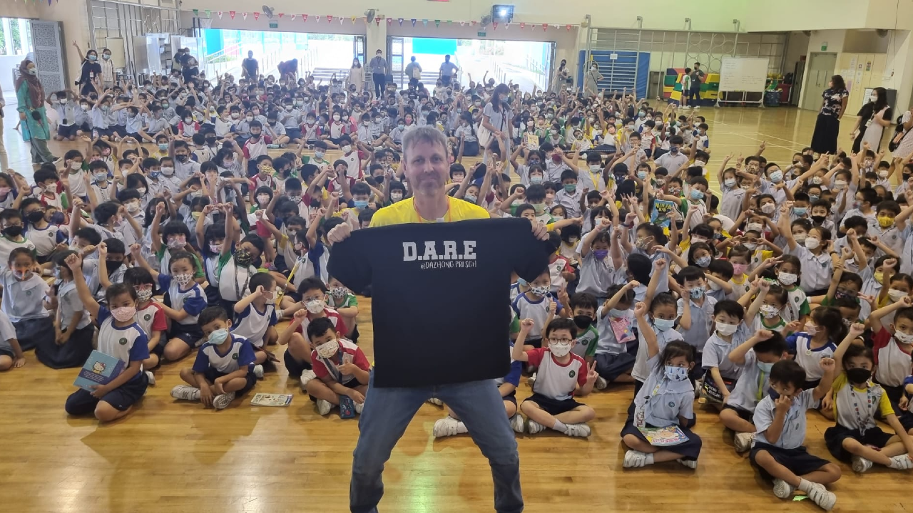 A man holds a black D.A.R.E. shirt in front of hundreds of masked schoolchildren seated in a gymnasium.