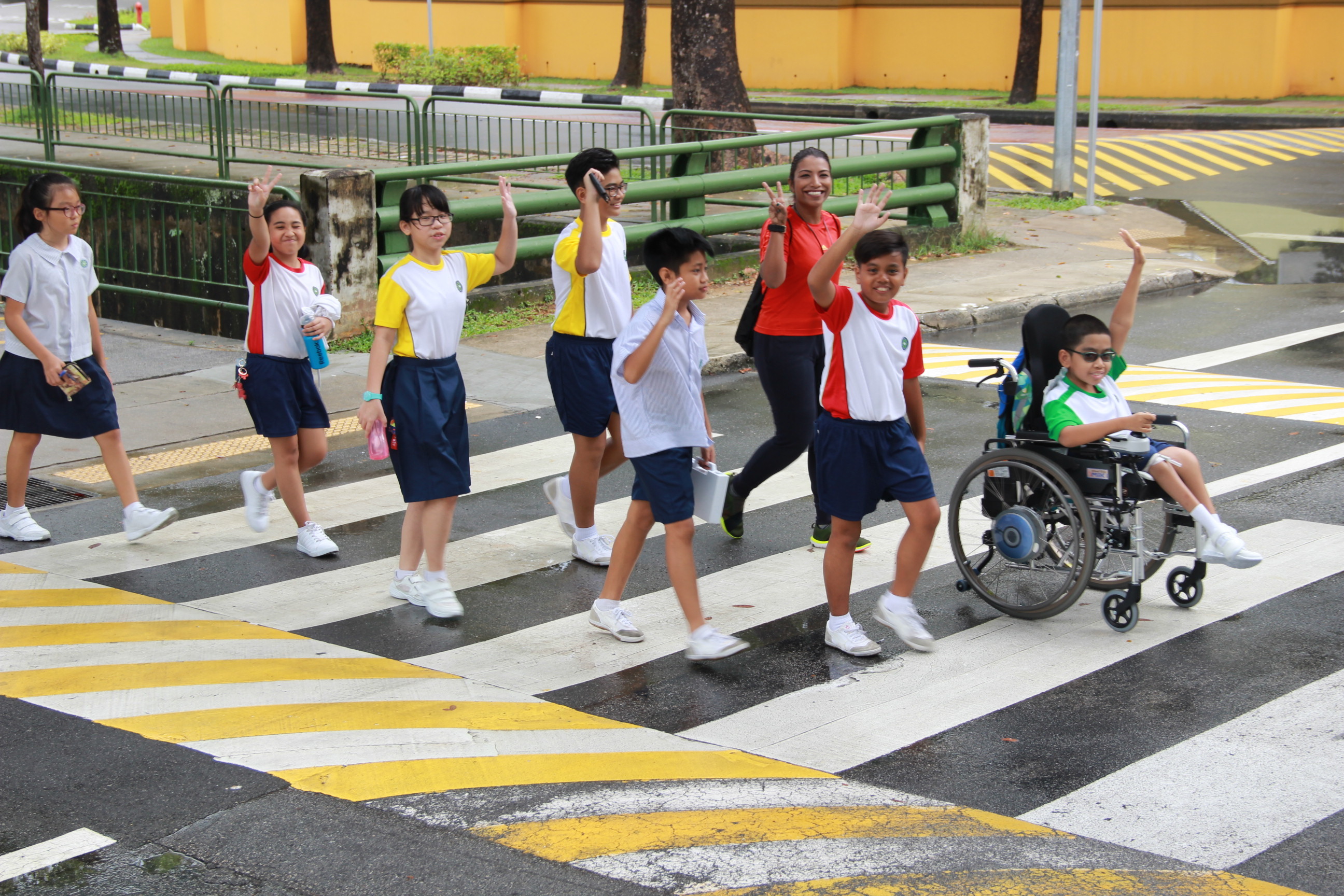 A group of school children and an adult cross a zebra crossing, one child in a wheelchair waves cheerfully.