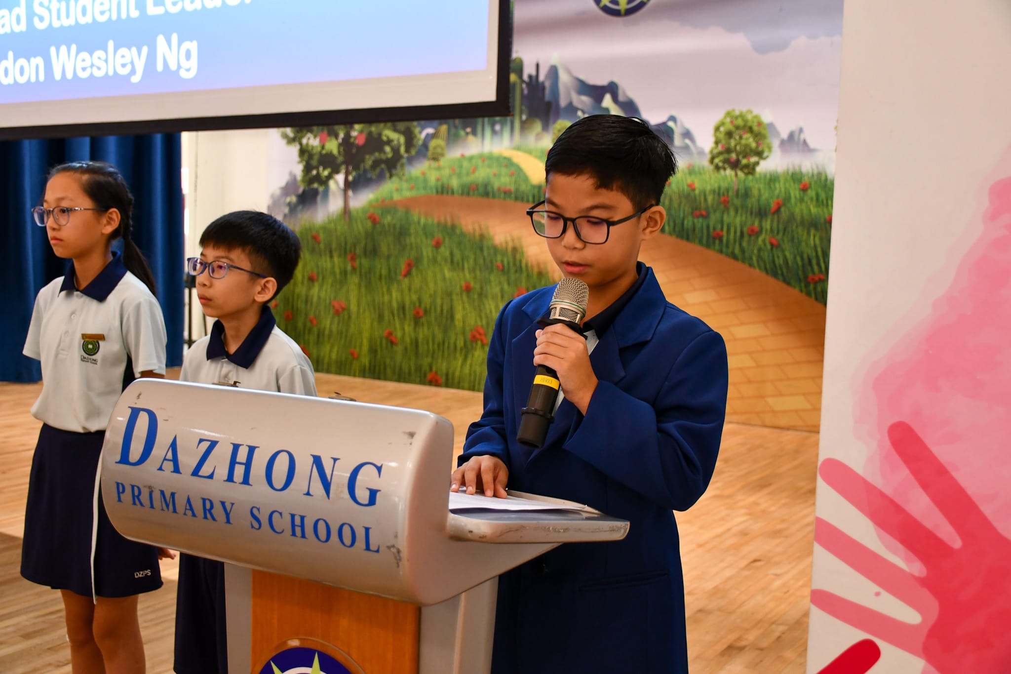 A boy in a blue blazer speaks at a Dazhong Primary School podium while two students in uniform stand behind him.
