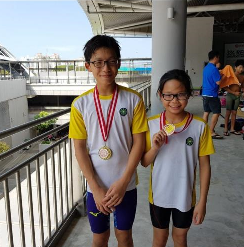 Two young swimmers wearing matching white and yellow jerseys pose with gold medals outdoors near a railing.