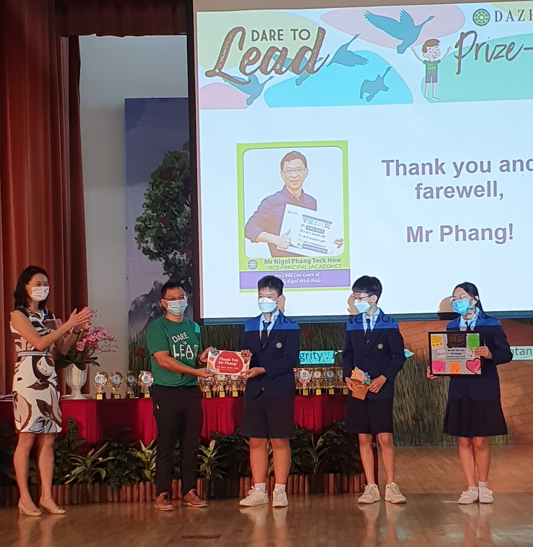 Students in uniforms present gifts to a man on stage at a Dare to Lead prize ceremony with a farewell slide for Mr Phang.