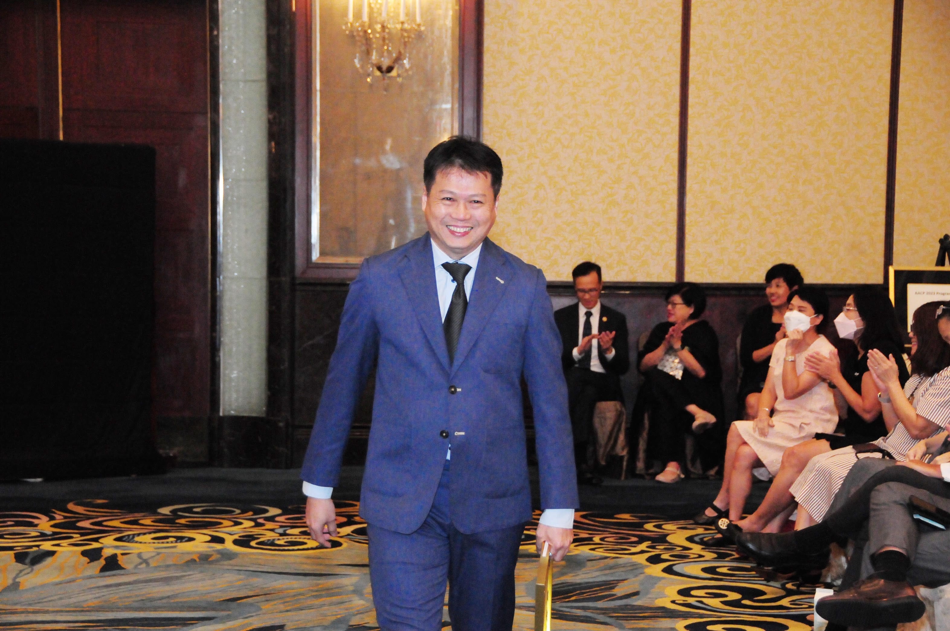 A smiling man in a blue suit walks through a formal ballroom as seated attendees applaud behind him.
