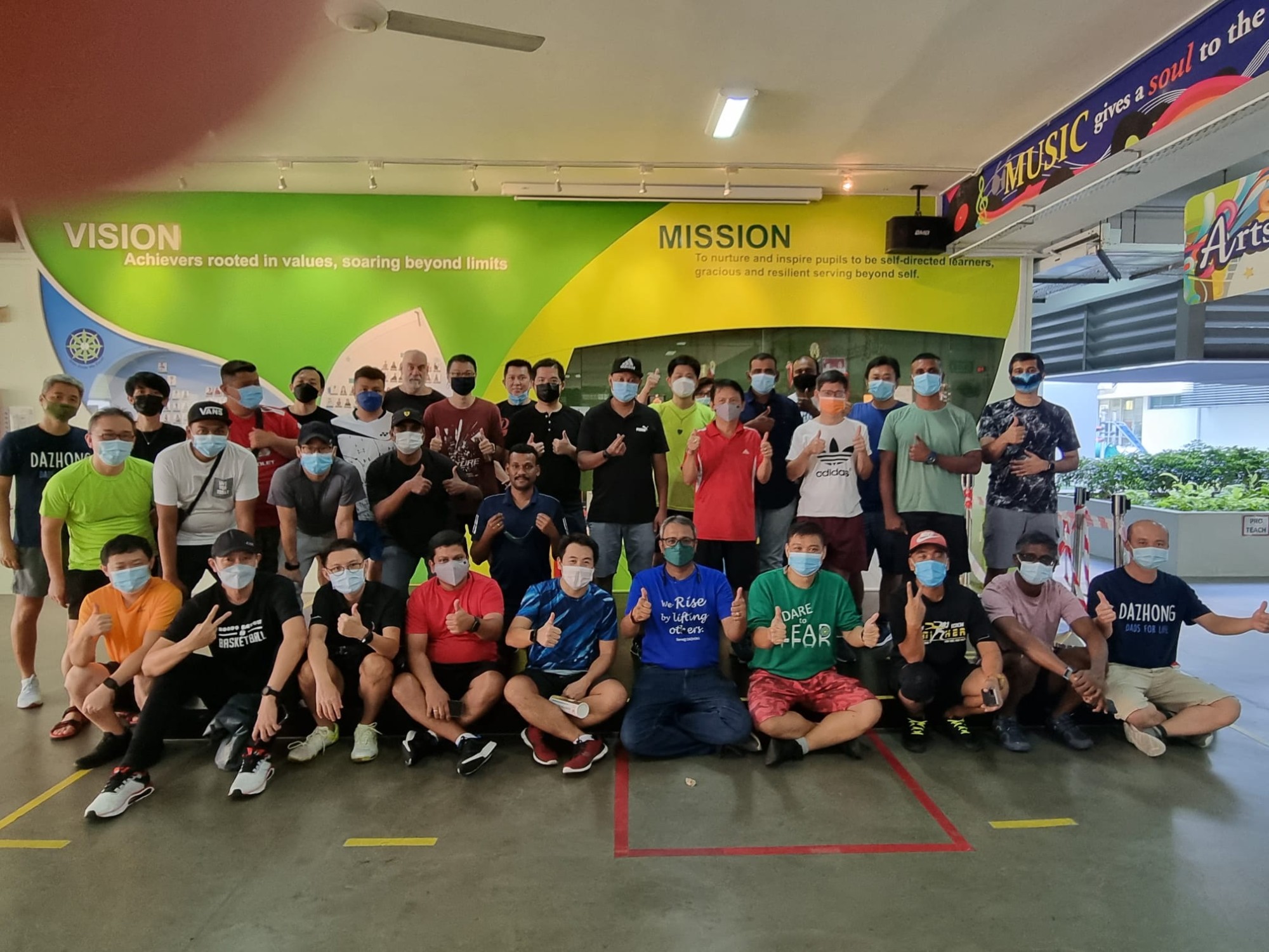 Large group of masked men posing with thumbs up in front of a school vision and mission wall mural.