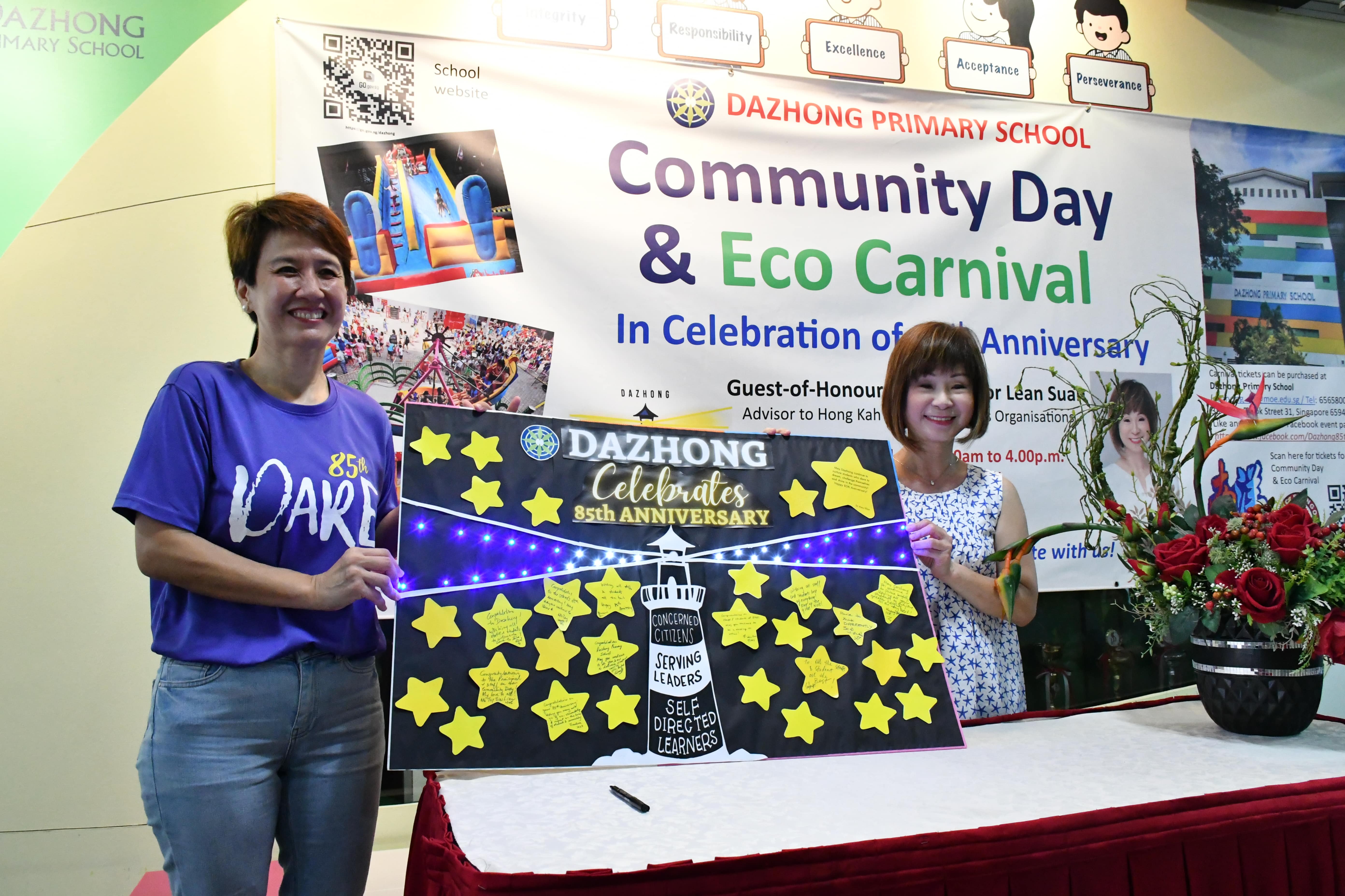 Two women hold a star-decorated board reading "Dazhong Celebrates 85th Anniversary" at a Community Day Eco Carnival.