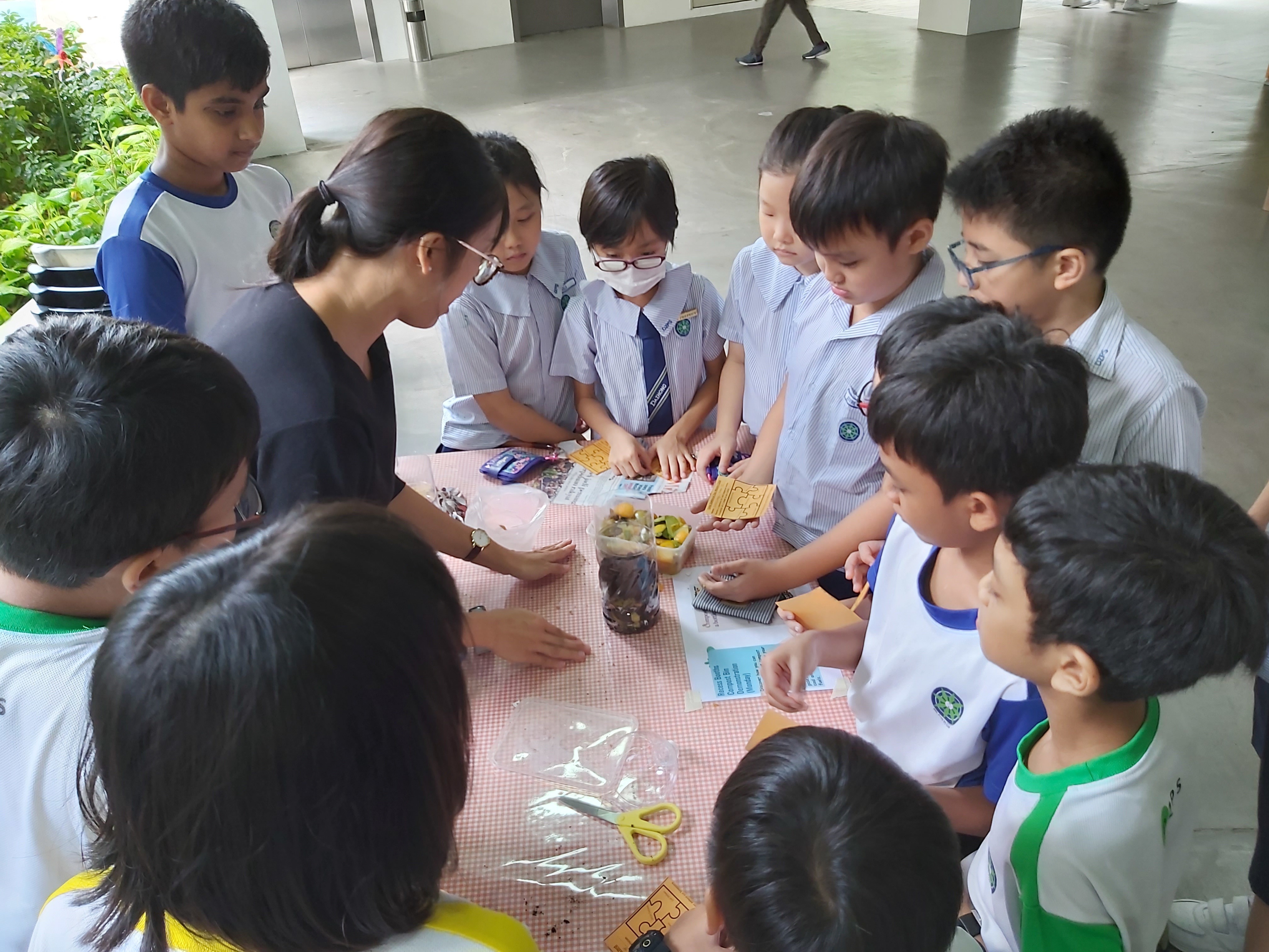 A teacher and uniformed schoolchildren gather around a table with soil, plants, scissors, and activity cards outdoors.