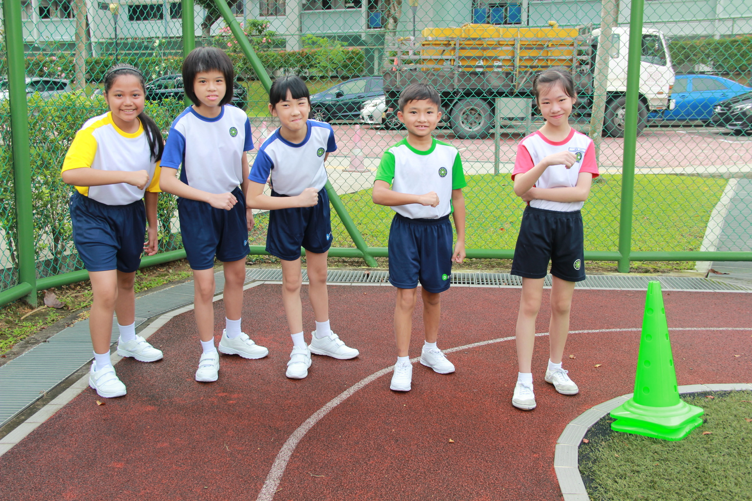 Five children in school PE uniforms stand on a red outdoor court, each making a fist gesture, with a green cone nearby.