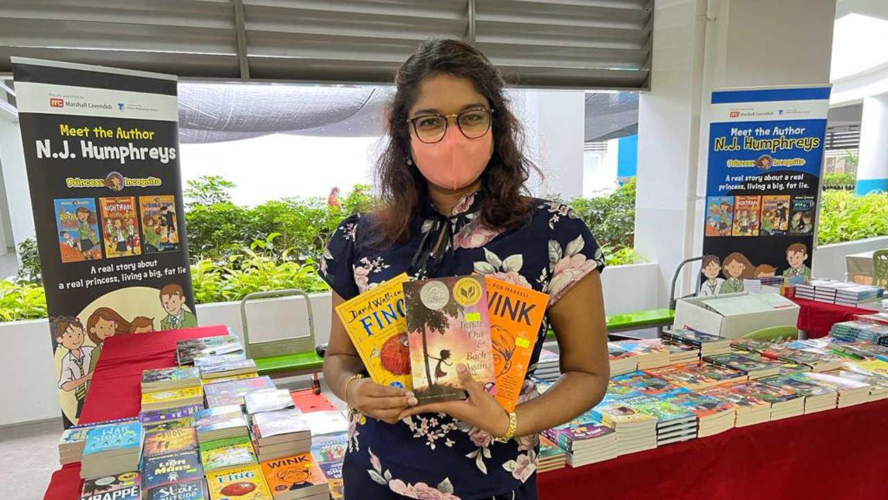 Woman wearing a mask holds three children's books at a book fair with N.J. Humphreys author event banners behind her.