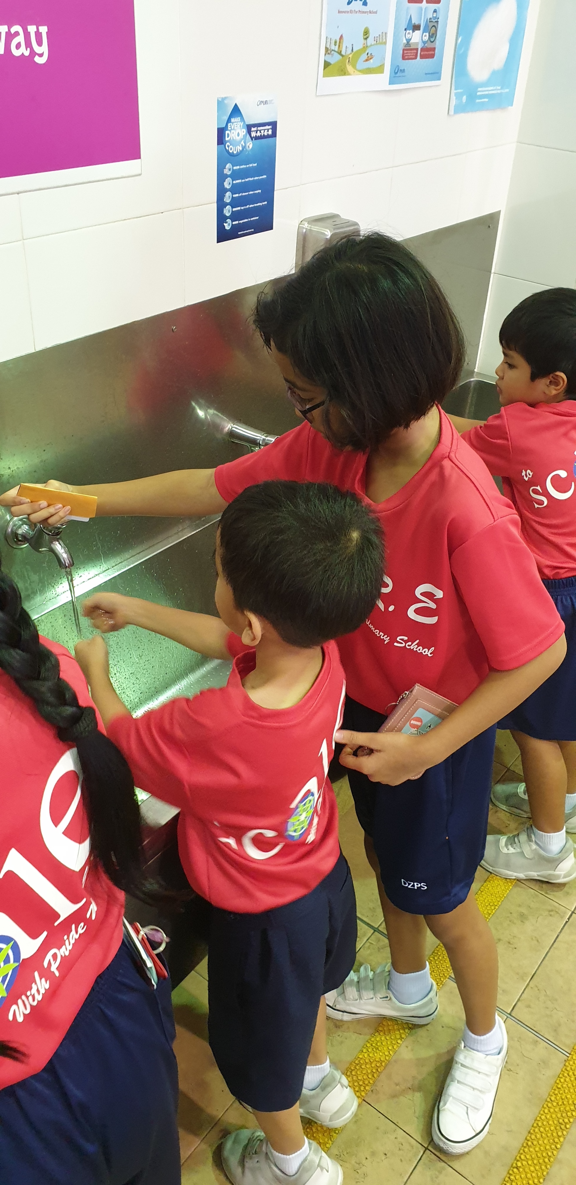 Children in red school uniforms wash hands at a metal sink, guided by an adult holding a bar of soap.