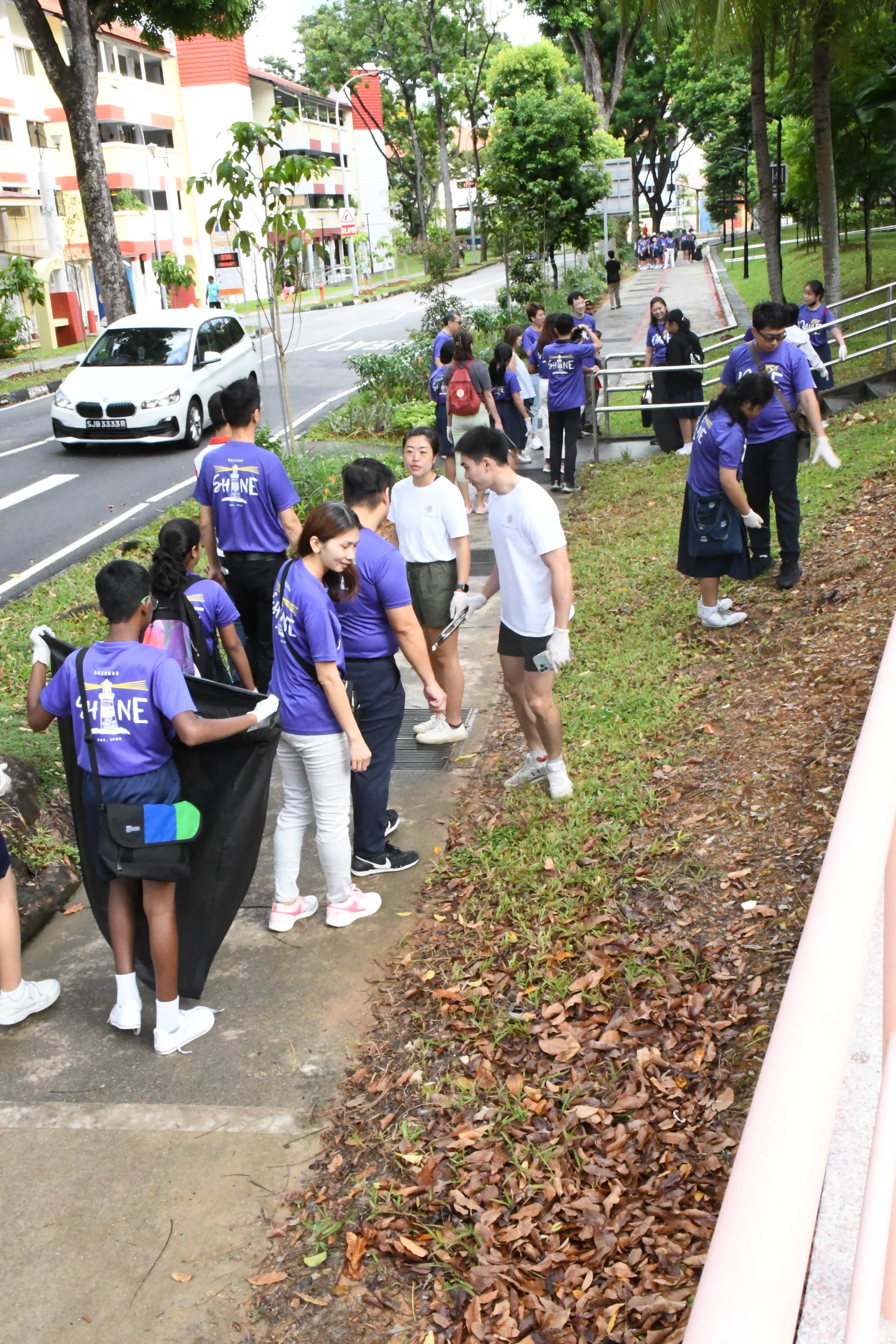 A large group in purple shirts collects litter along a residential street sidewalk with fallen leaves nearby.