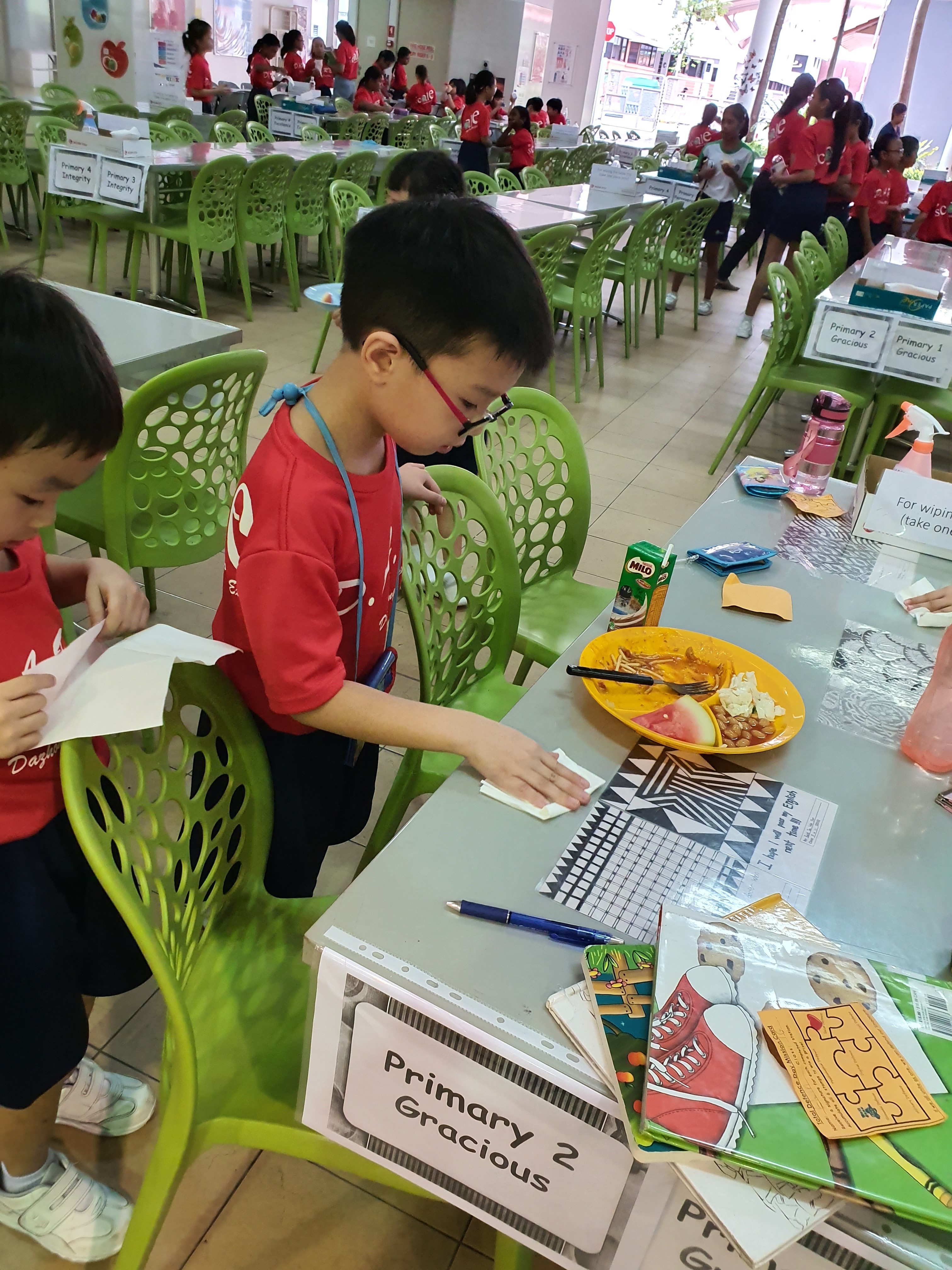 Young boy in red uniform wiping a cafeteria table with a sign reading Primary 2 Gracious, food on a yellow plate nearby.