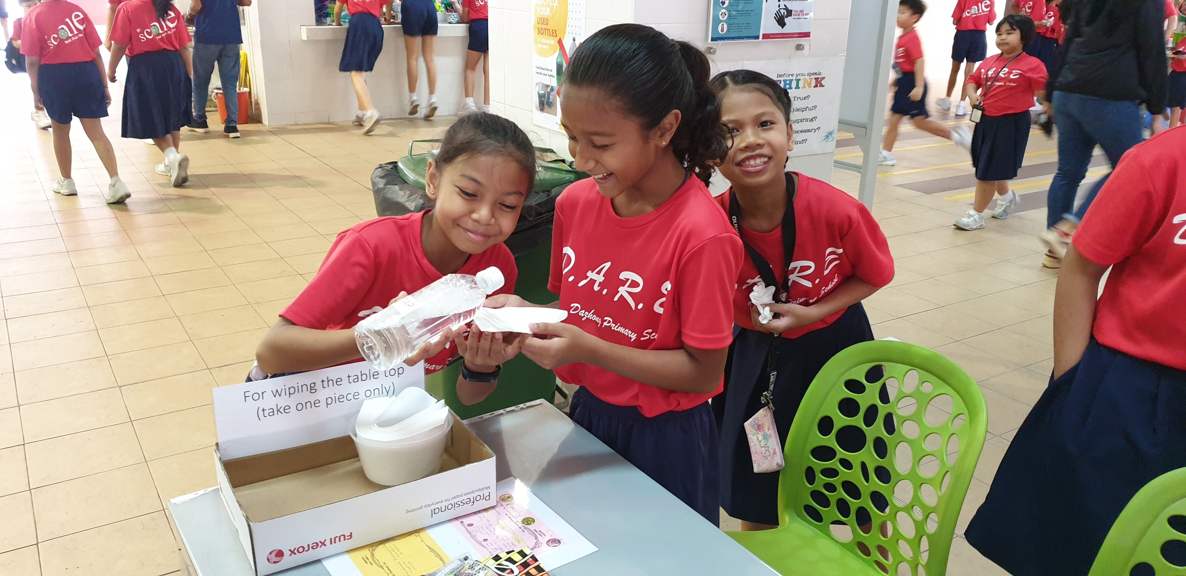 Three girls in red D.A.R.E. shirts smile while handling a water bottle at a school activity table.