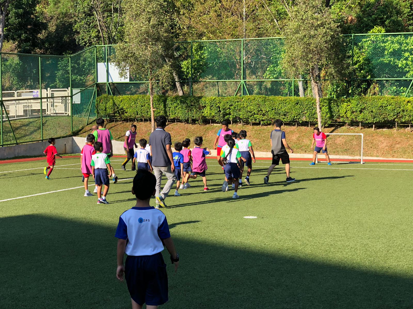 Children in colorful vests play soccer on a green turf field with a goal, supervised by an adult coach.