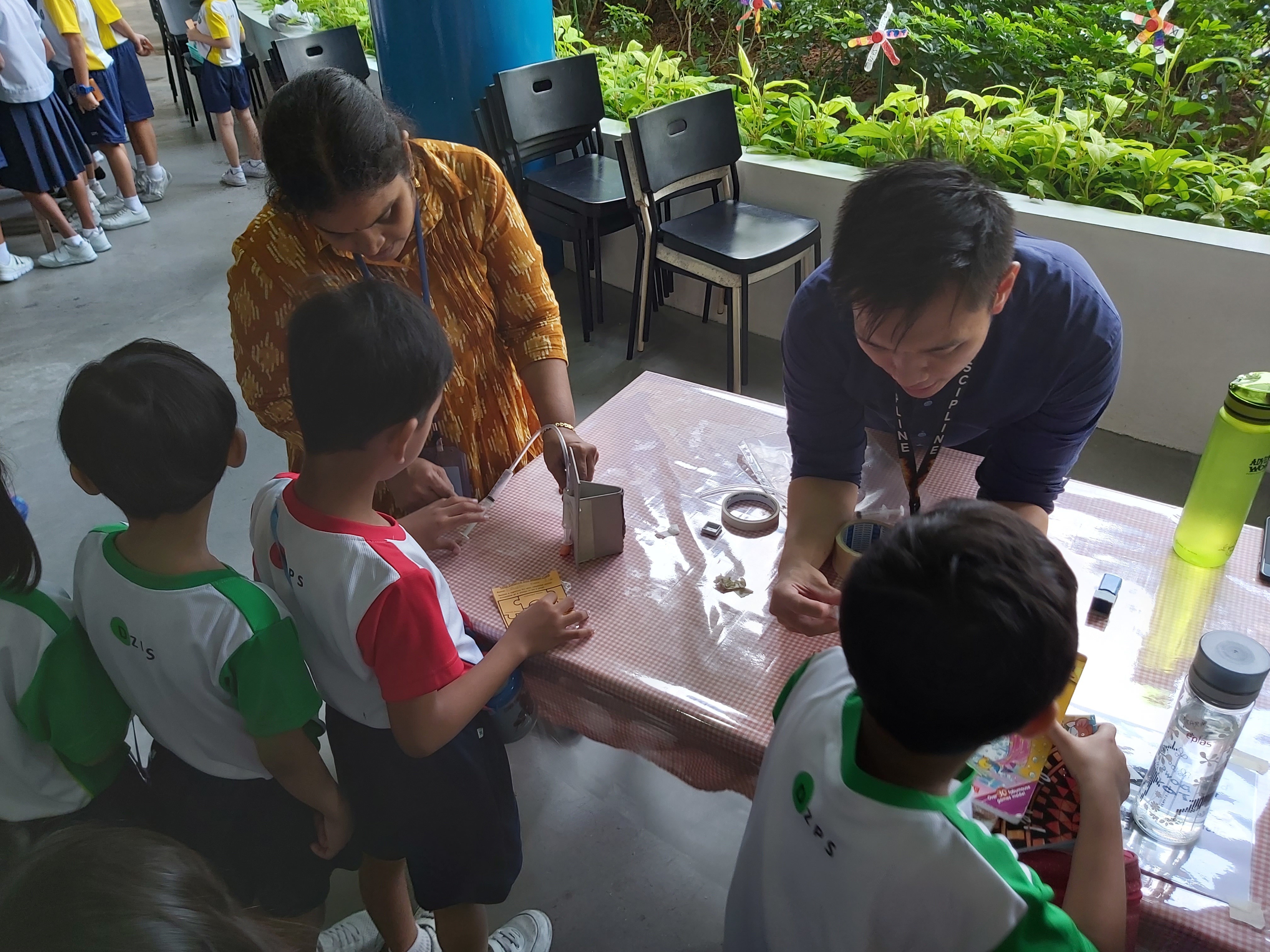 Two adults guide young children in a hands-on craft activity at a table outdoors near green plants.
