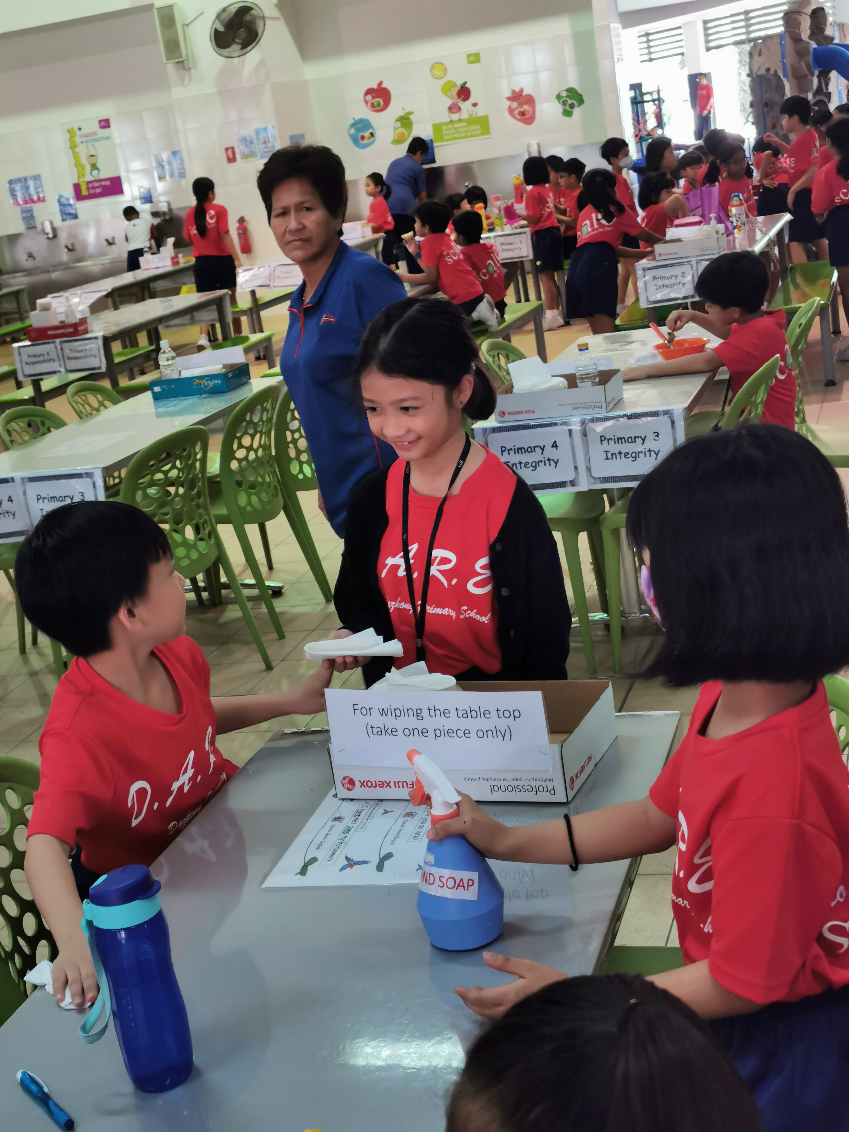 Children in red school shirts clean cafeteria tables using soap and wipes, supervised by an adult in a blue shirt.