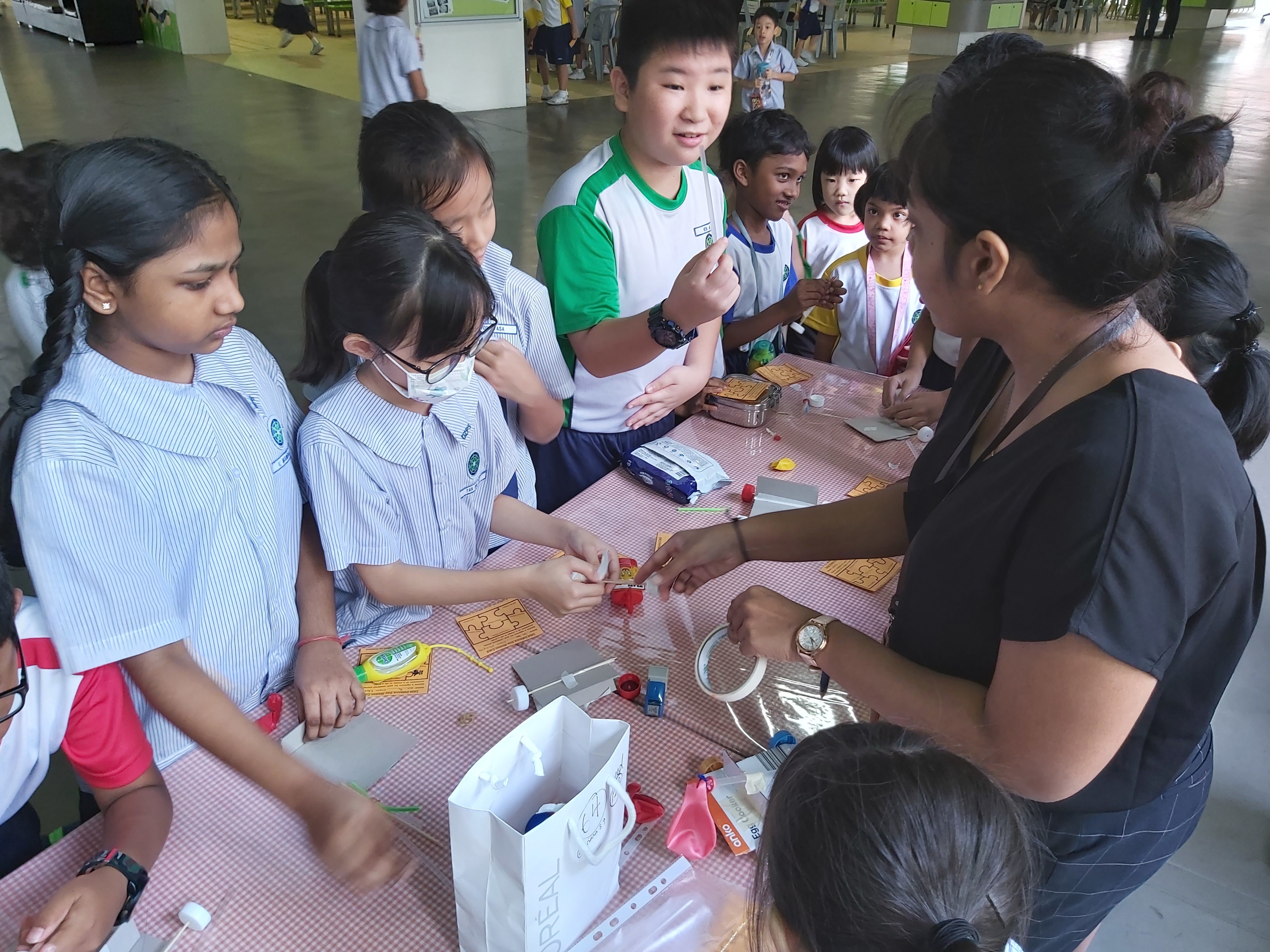 Several school children in uniforms gather around a table with craft supplies, guided by an adult instructor.