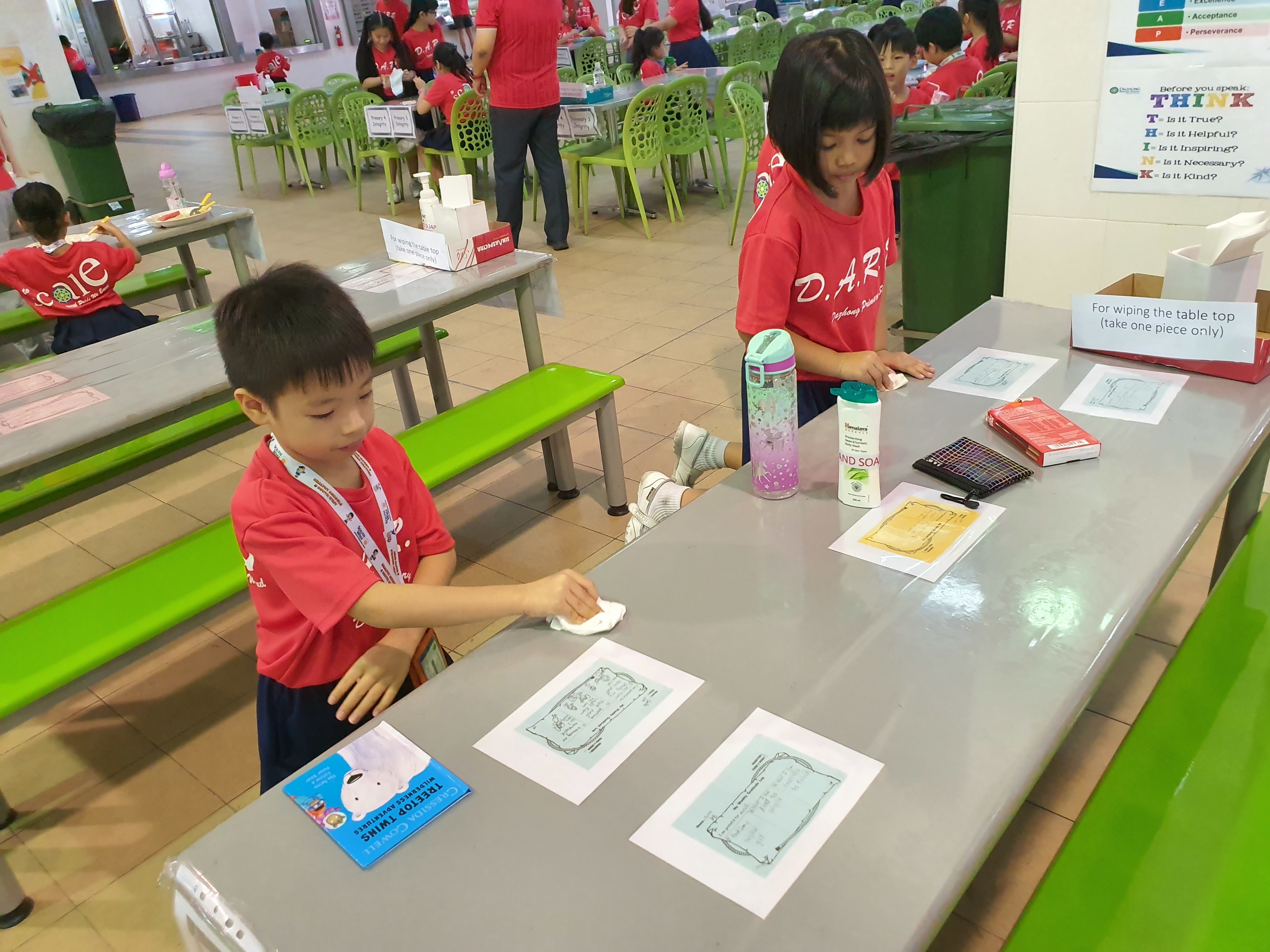 Two children in red shirts wipe a cafeteria table with papers and supplies spread on it.