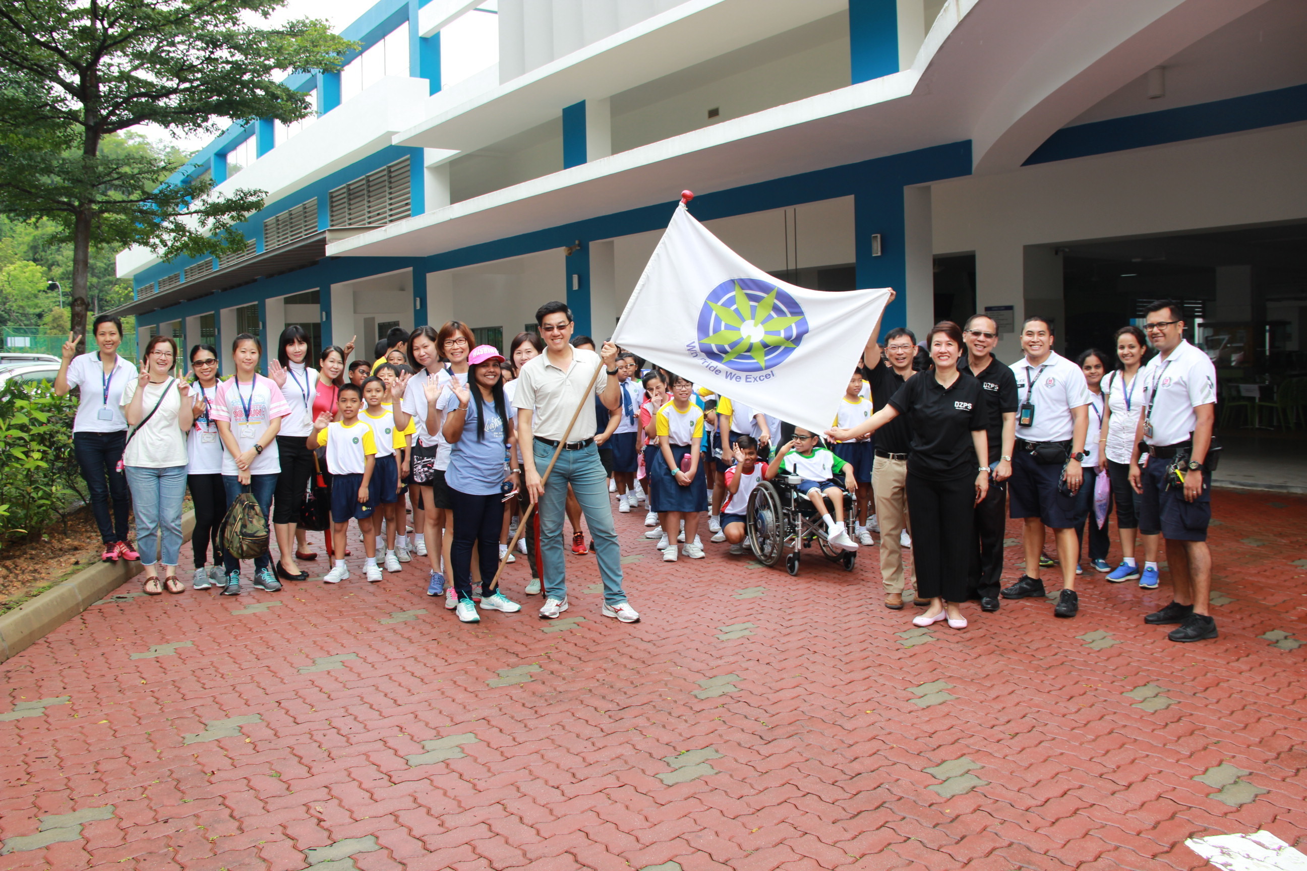 A large group of adults and children pose outside a school building, with a man holding a white flag bearing a blue emblem.