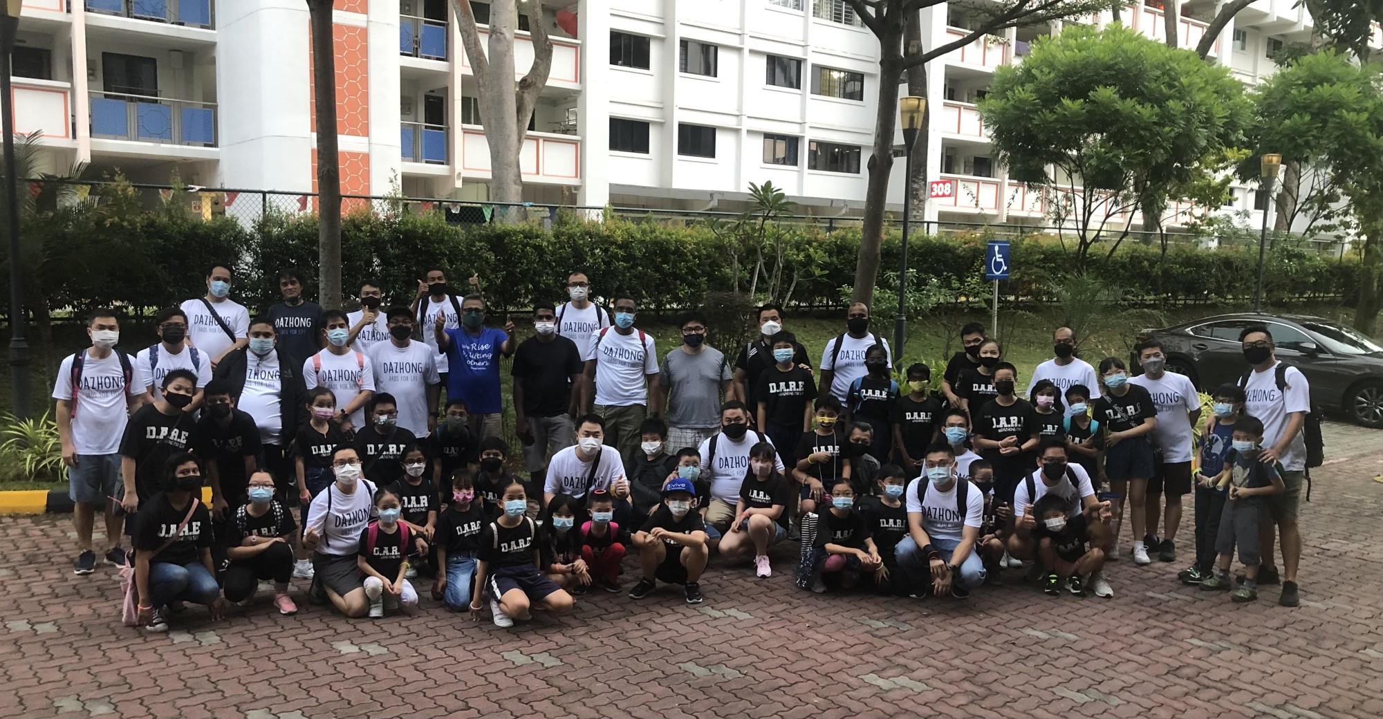Large group of masked adults and children wearing Dazhong and D.A.R.E shirts posing outdoors near apartment buildings.