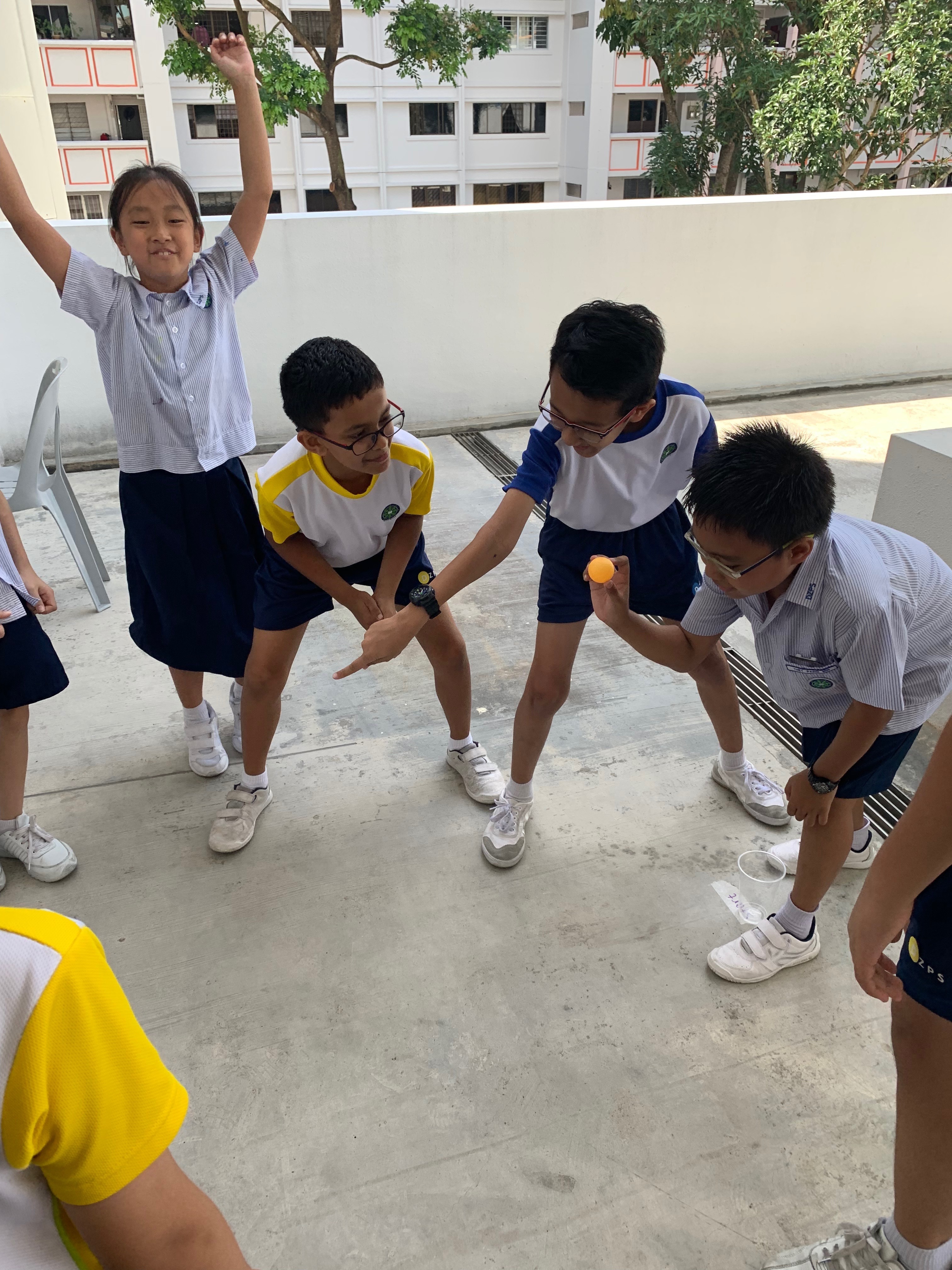 Four school children in uniforms play outdoors; one holds an orange ball while a girl raises her arms in celebration.