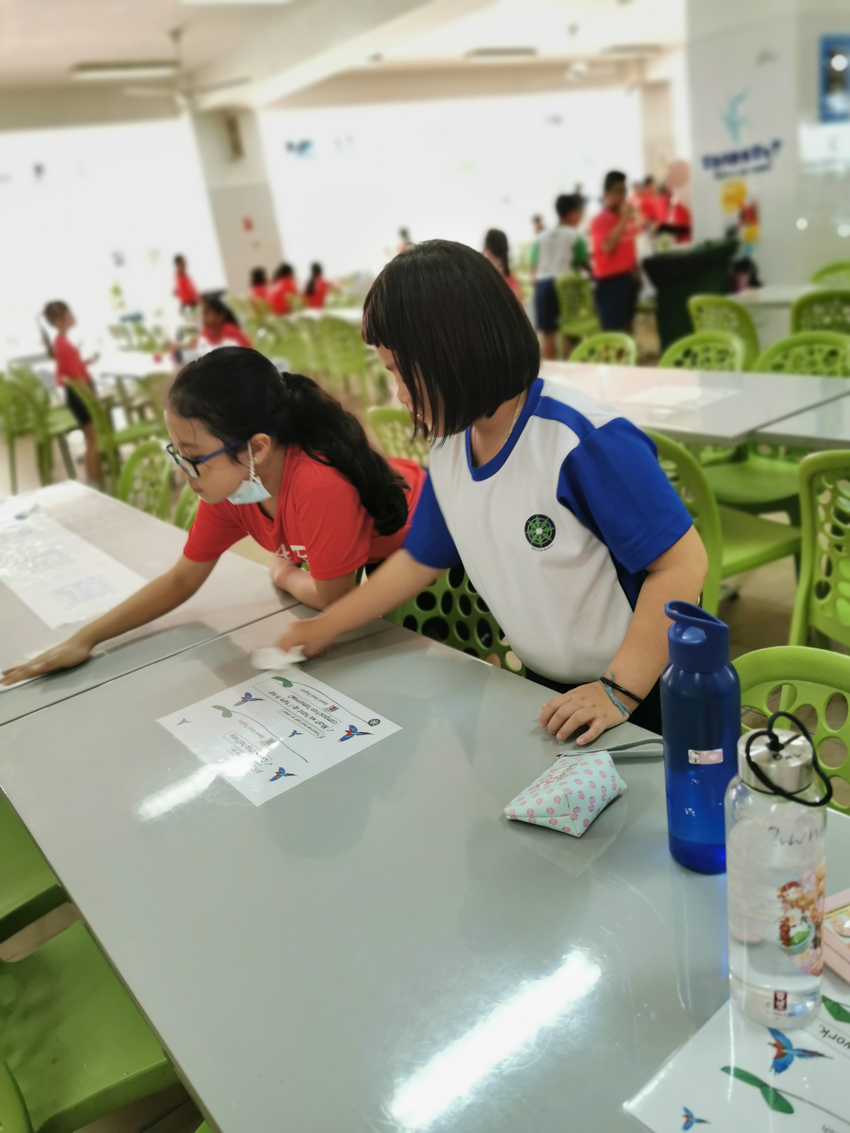 Two students wipe down a cafeteria table, with green chairs and other students in the background.