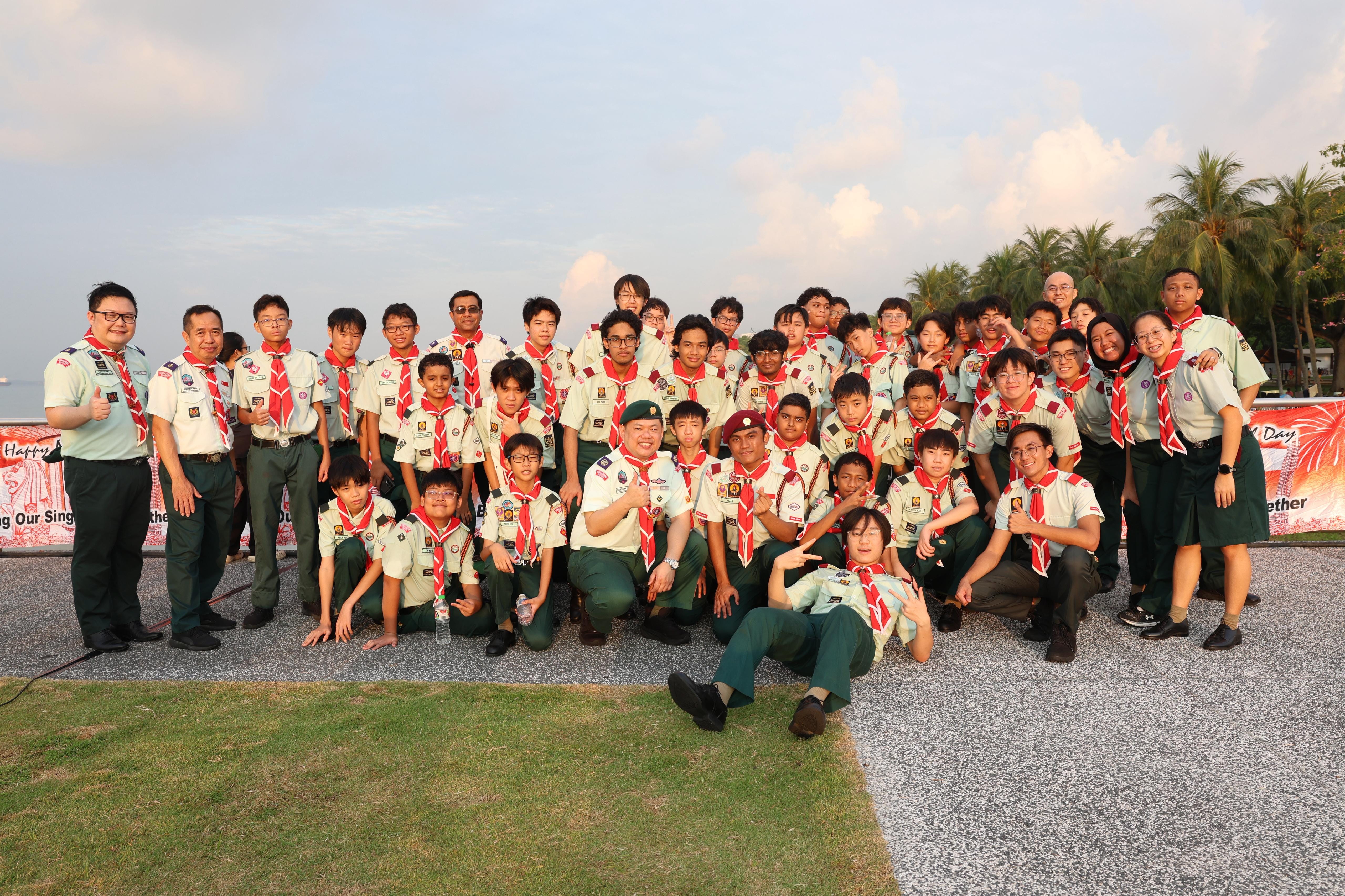 Large group of scouts in green uniforms with red neckerchiefs posing outdoors near palm trees and a coastal area.
