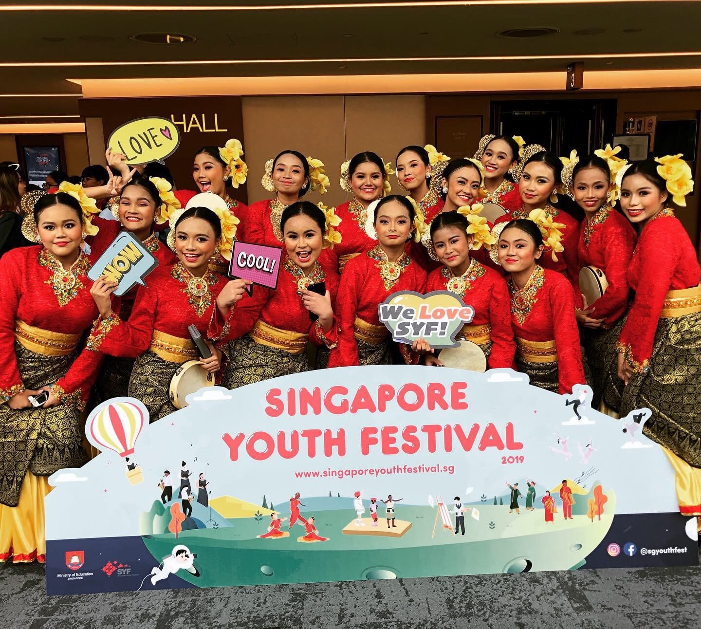 Group of young women in red and gold traditional malay costumes posing with the Singapore Youth Festival 2019 sign.