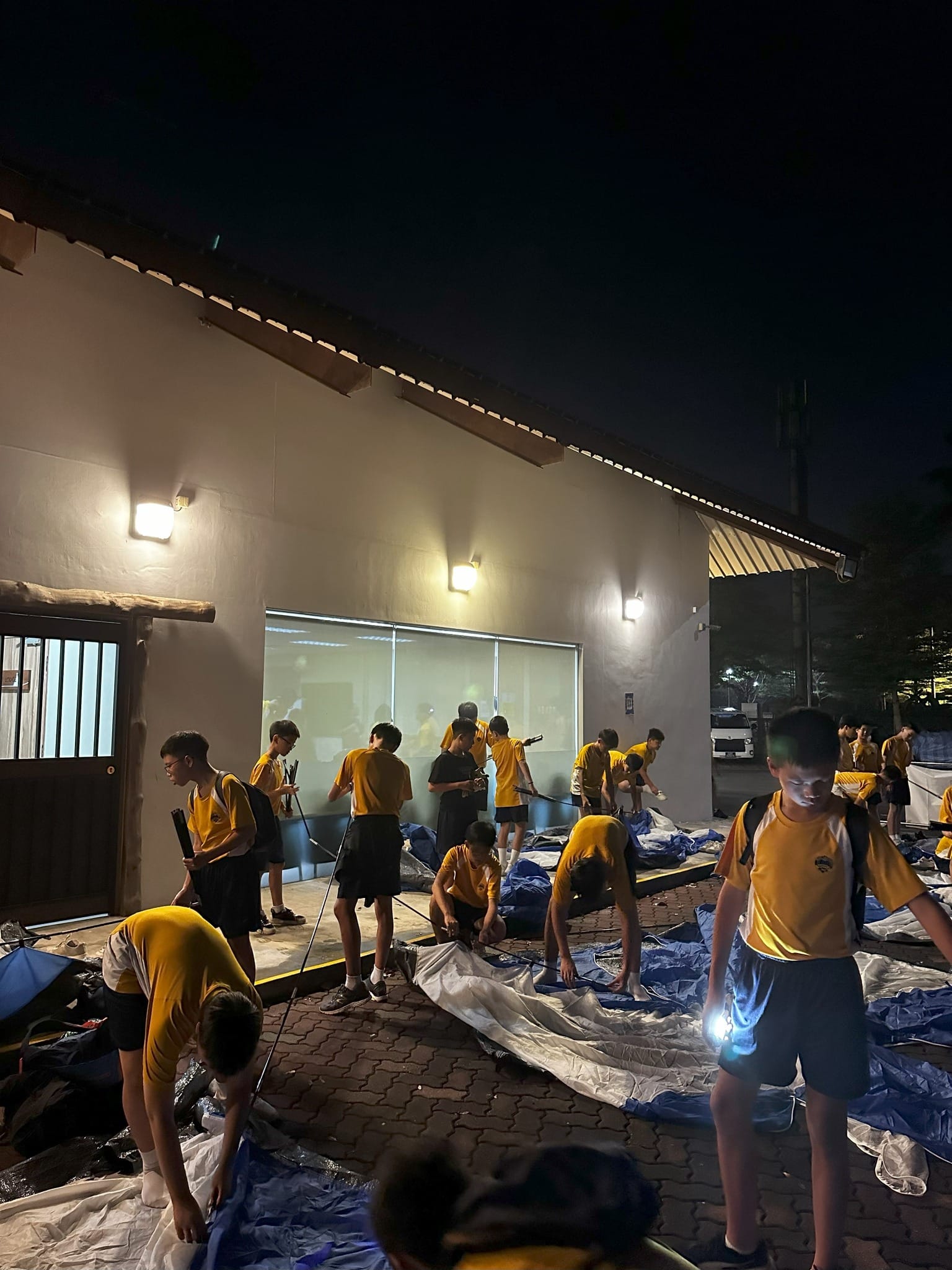 nighttime scene outside a brightly lit camp building where Secondary One students work in teams to pitch their blue and white camping tents on a brick-paved ground. The students, wearing their yellow-and-white school sports uniforms, bend down to lay out the tent fabric and assemble the structural poles. In the foreground, one student uses a flashlight to illuminate the ground for his group. The building's exterior wall is illuminated by three bright outdoor lamps under a dark night sky.