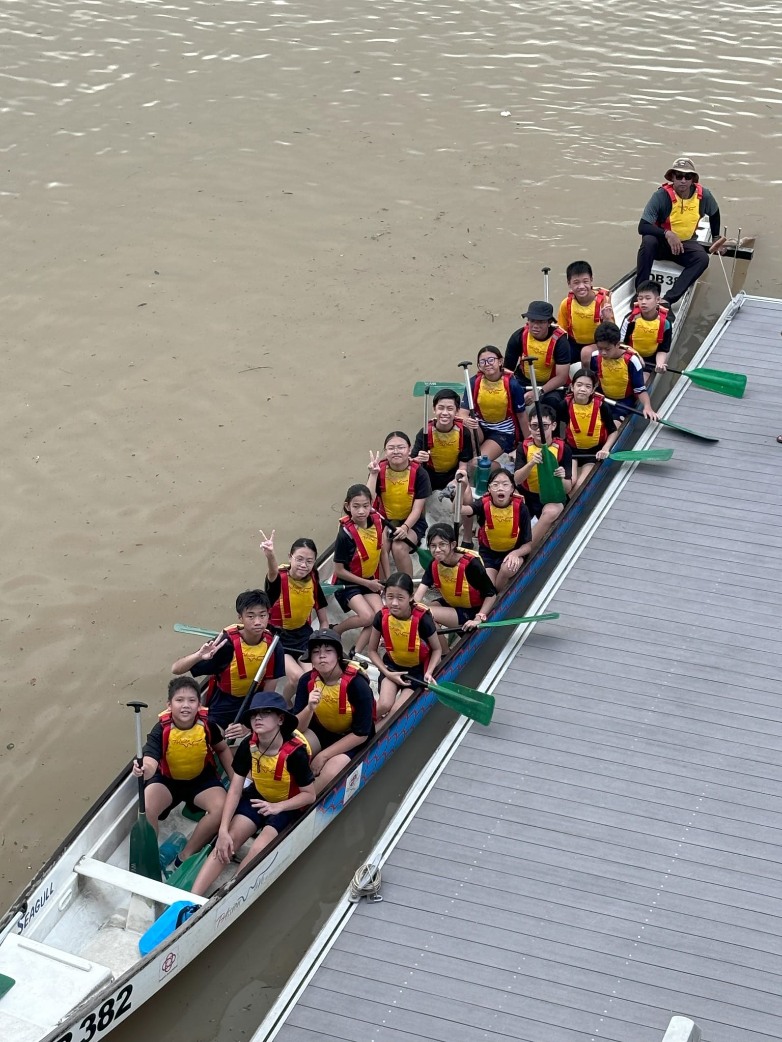 A group of young paddlers in yellow life vests sit in a long dragon boat beside a dock on a murky river.