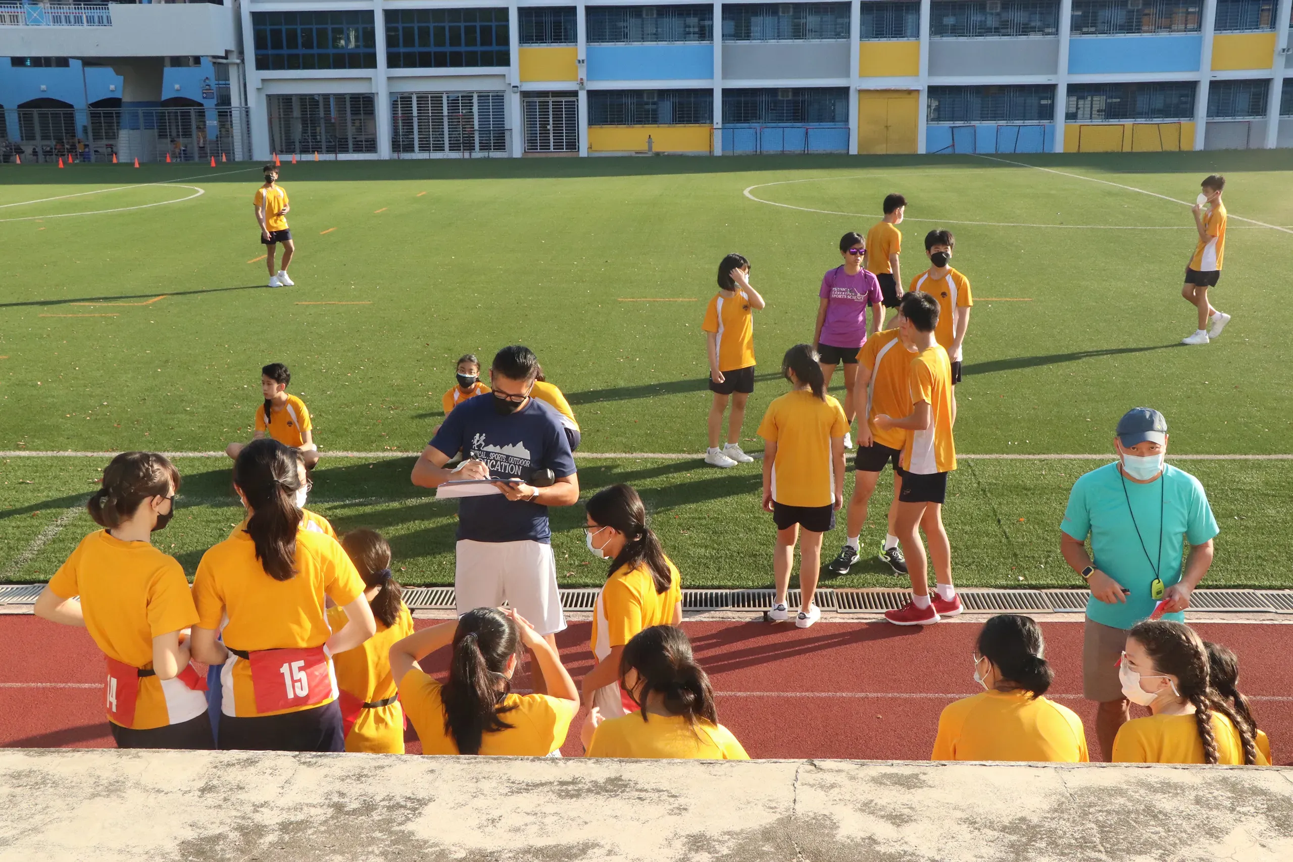 PE teachers facilitating and briefing students on the running track and artificial turf field during the Fairfield Methodist School (Secondary) Annual Run in 2022. Students are wearing yellow and purple sports attire and face masks.