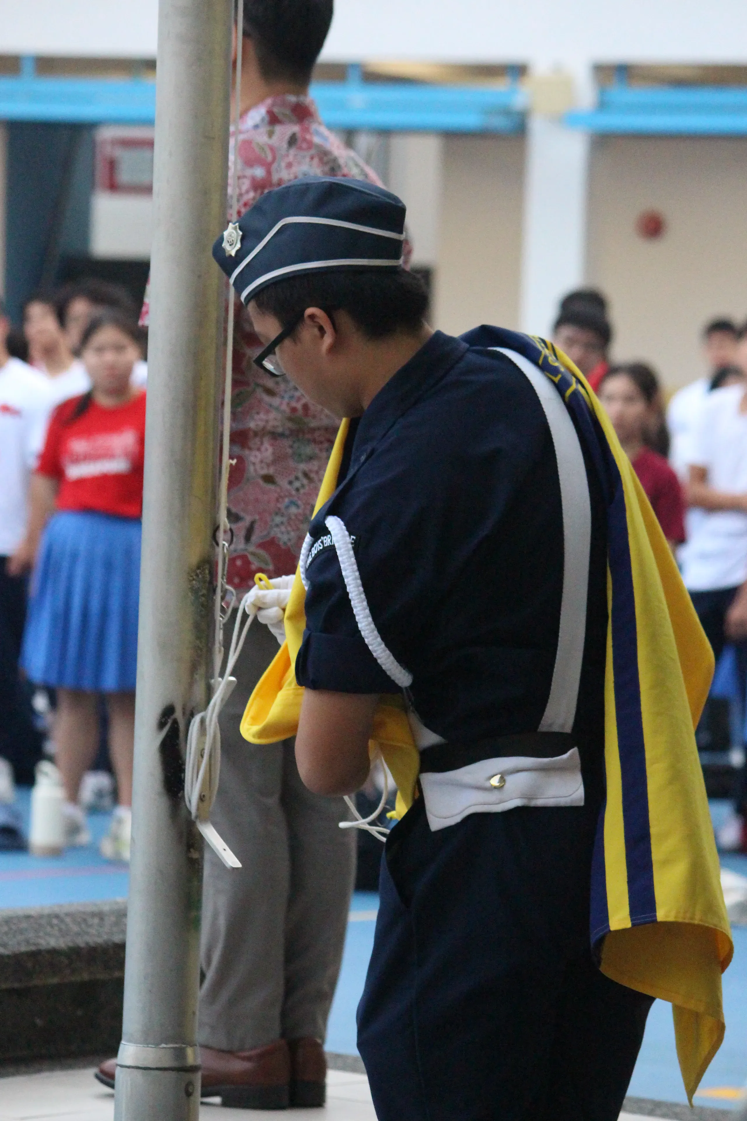 A close-up, side profile view of a Boys' Brigade cadet preparing a flag for the flag-raising ceremony in the parade square during National Day celebrations. The cadet is wearing a dark blue uniform with white lanyards, white gloves, and a formal peak cap. A folded yellow and blue school flag is draped over his left shoulder as he focuses on securing the clips to the flagpole's halyard rope. In the blurred background, Principal Mr. Wee and students stand at attention.