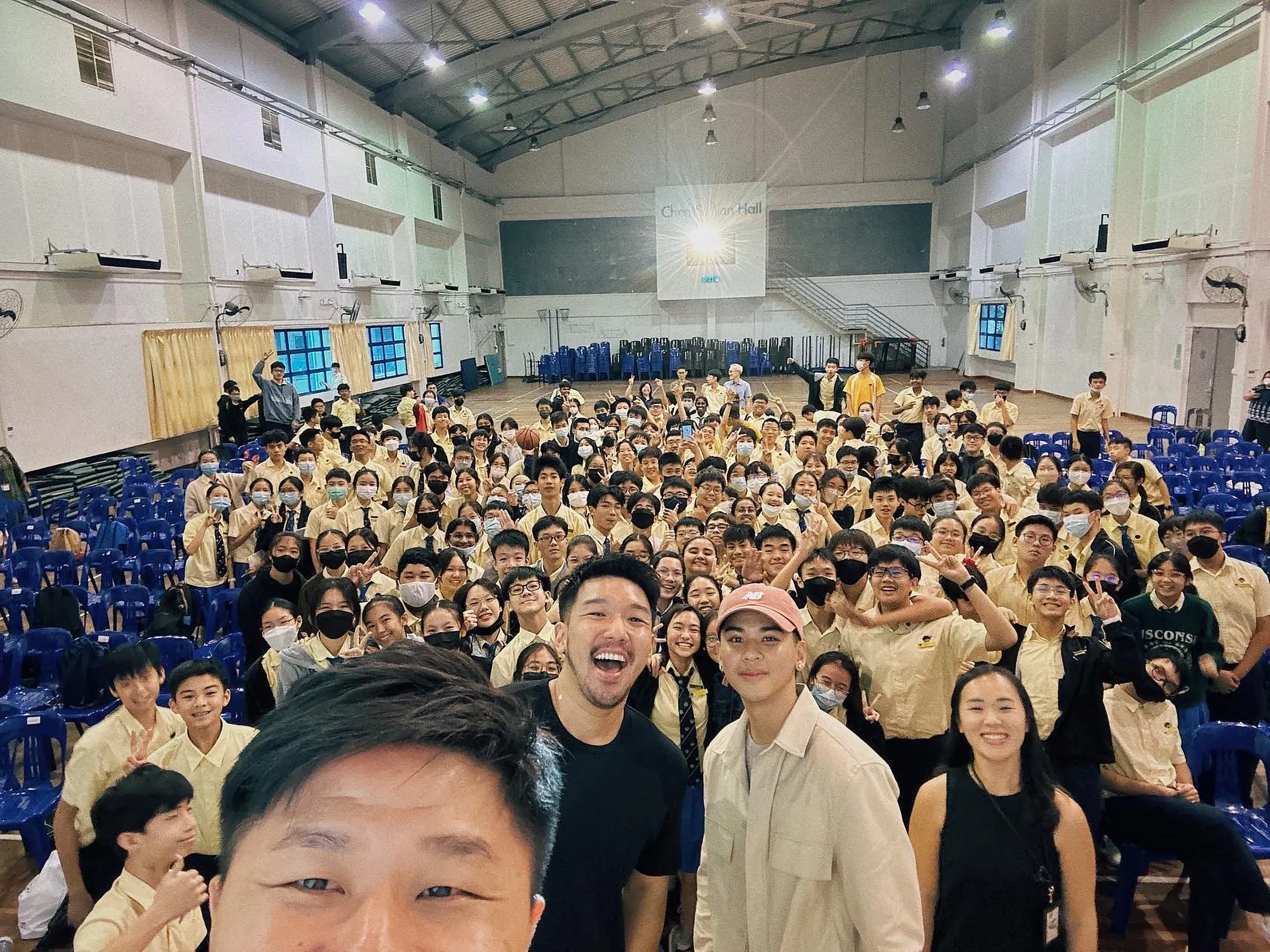 A large group selfie featuring Secondary 3 students in their cream school uniforms gathered inside the school hall (Chen Su Lan Hall). Several guest speakers and teachers smile broadly in the foreground, while a large crowd of enthusiastic students wave and pose on the tiered floor seating behind them.
