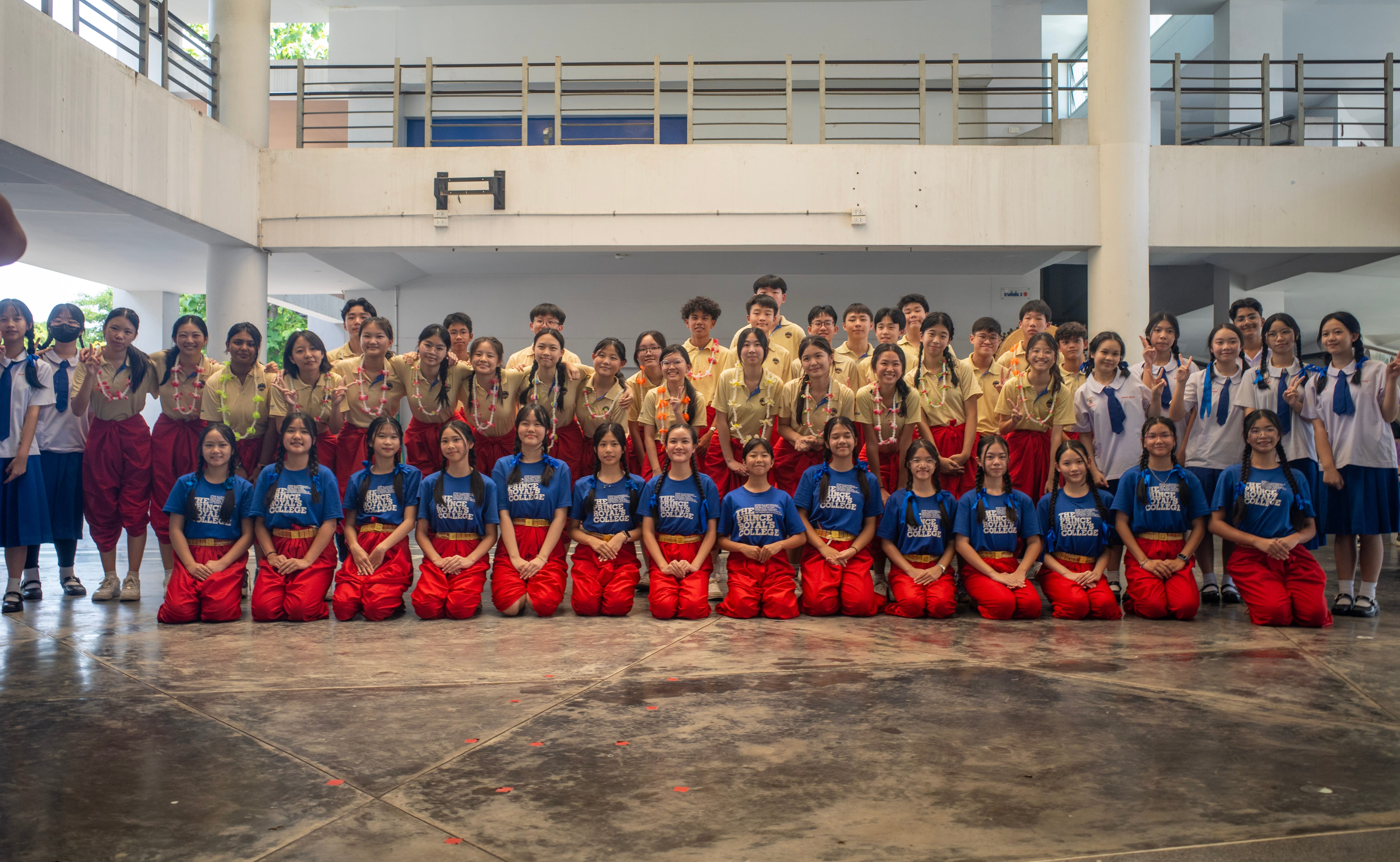 A large group of secondary school students and their hosts pose for a commemorative group photo inside an open-air school courtyard with a multi-tiered balcony layout. In the front row, a line of female students kneel on the polished floor wearing bright blue "The Prince Royal's College" t-shirts and traditional Thai red chong kraben trousers with gold belts. Standing behind them is a large group of students wearing light yellow school shirts, some adorned with decorative floral garlands (lei) around their necks. They are flanked on both ends by peers in white and blue Thai school uniforms, all smiling together to celebrate the cultural exchange.