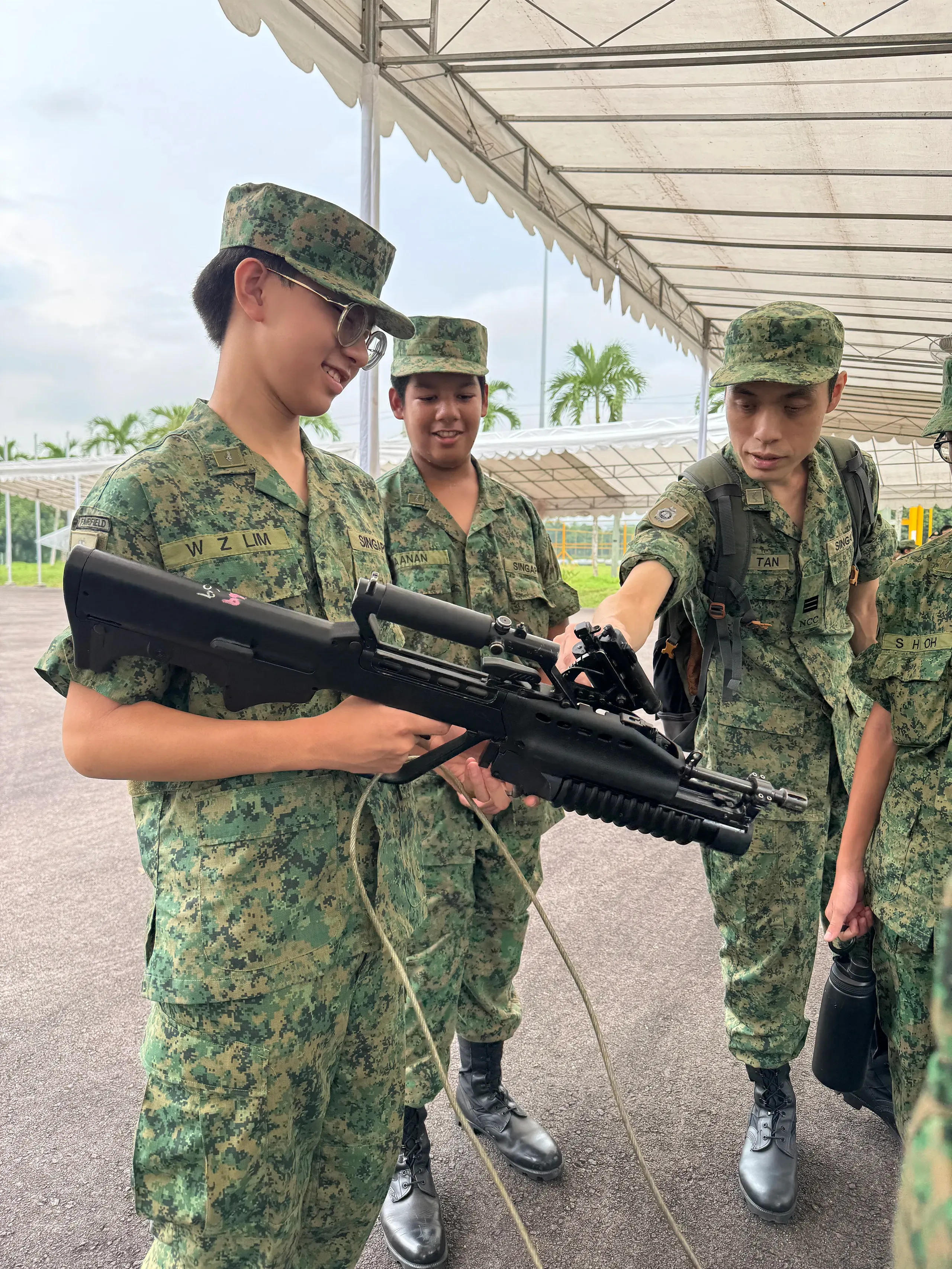A Fairfield teachers in NCC uniform guides a Fairfield NCC cadet in a green pixelated camouflage uniform on how to handle a training rifle under an outdoor tent structure, while another cadet watches closely in the background.