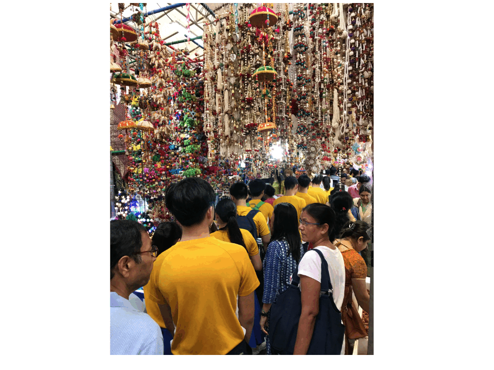 Crowd of people walking through a colorful market stall with hundreds of hanging decorations and trinkets overhead.