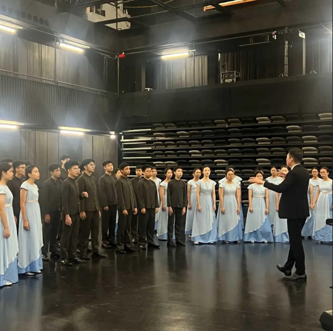 Members of the school choir are gathered on a dark, reflective stage for a vocal warm-up session before their SYF performance. The female choristers stand on the left and right flanks wearing long, light blue formal gowns, while the male choristers are grouped in the center wearing high-collared black shirts and dark trousers. On the right, the choir instructor stands in profile, dressed in a sharp black suit, with his hands raised to guide the students' breathing and pitch. The background features high studio ceilings, professional stage lighting rigs, and stacked rows of auditorium seating.