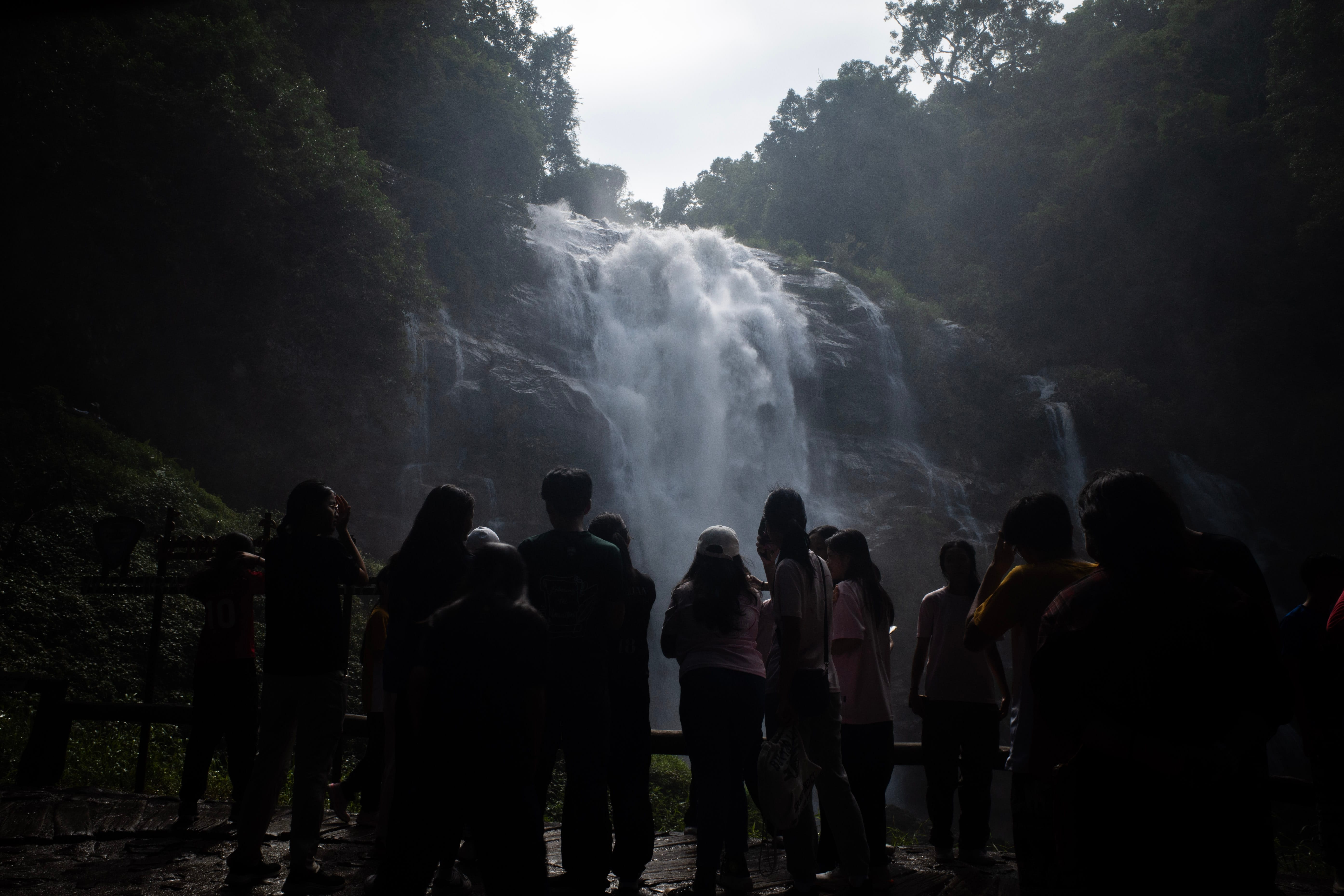 A group of secondary school students are seen from a back-facing silhouette view as they admire a massive, powerful waterfall cascading down a rocky cliffside in Chiang Mai during their TIE trip. The bright sunlight catches the heavy white mist and spray rising from the rushing water, creating a stark contrast against the dark silhouettes of the students standing on a wooden viewing platform in the foreground. Lush, dense green jungle foliage lines the sides and top of the waterfall under an overcast sky.