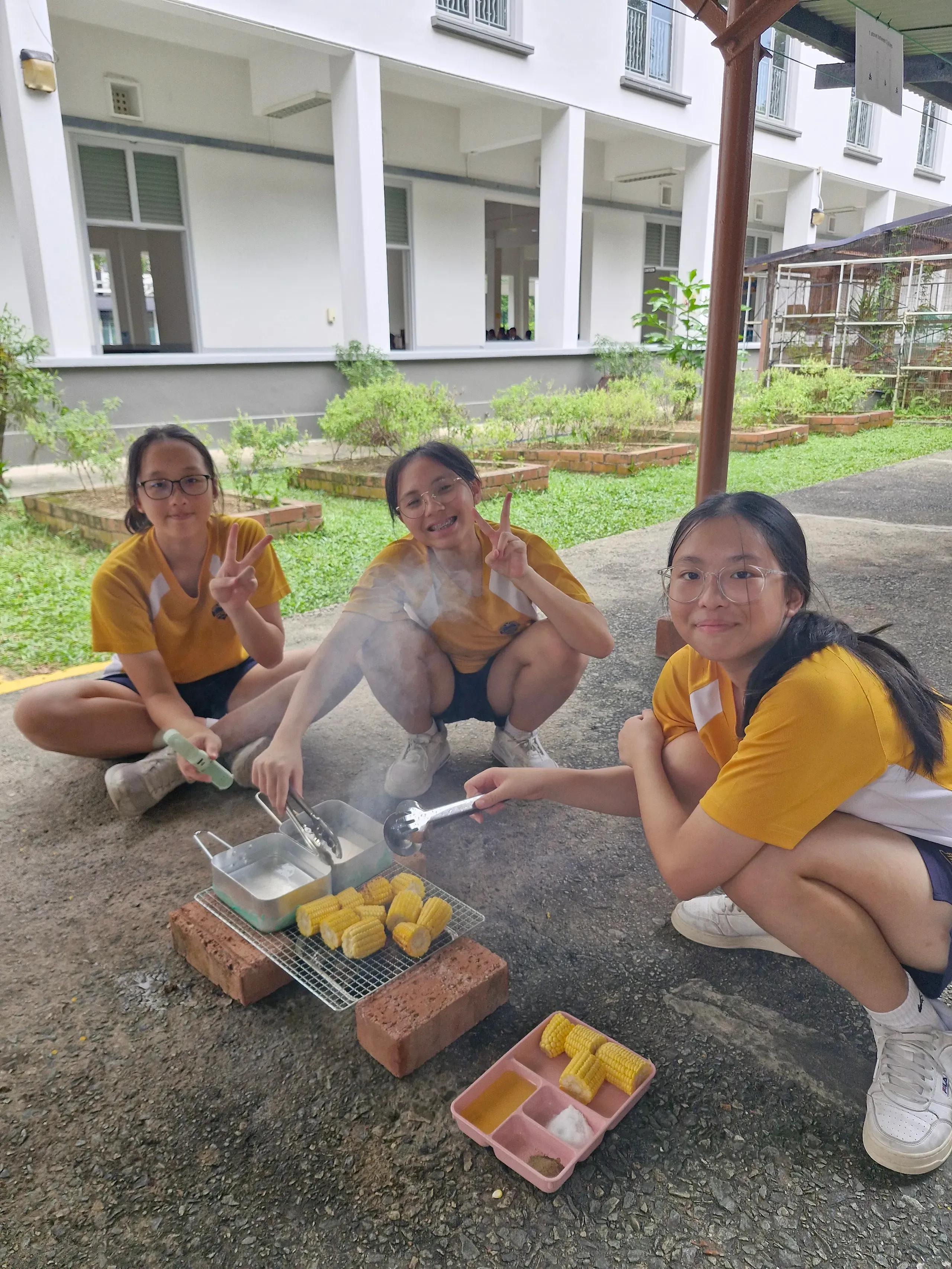 Three female student leaders smiling and posing for a photo while cooking corn on the cob over an outdoor brick-and-charcoal grill during their training camp. Seated and squatting on the ground, the students wear yellow and white school sports t-shirts and dark shorts. The two students on the left make peace signs at the camera, while the student on the right uses tongs to adjust the corn pieces on the wire mesh grill next to two metal mess tins. A plastic tray containing more corn pieces and ingredients sits on the ground in the foreground. In the background, an open grassy courtyard and a white multi-story school building are visible.