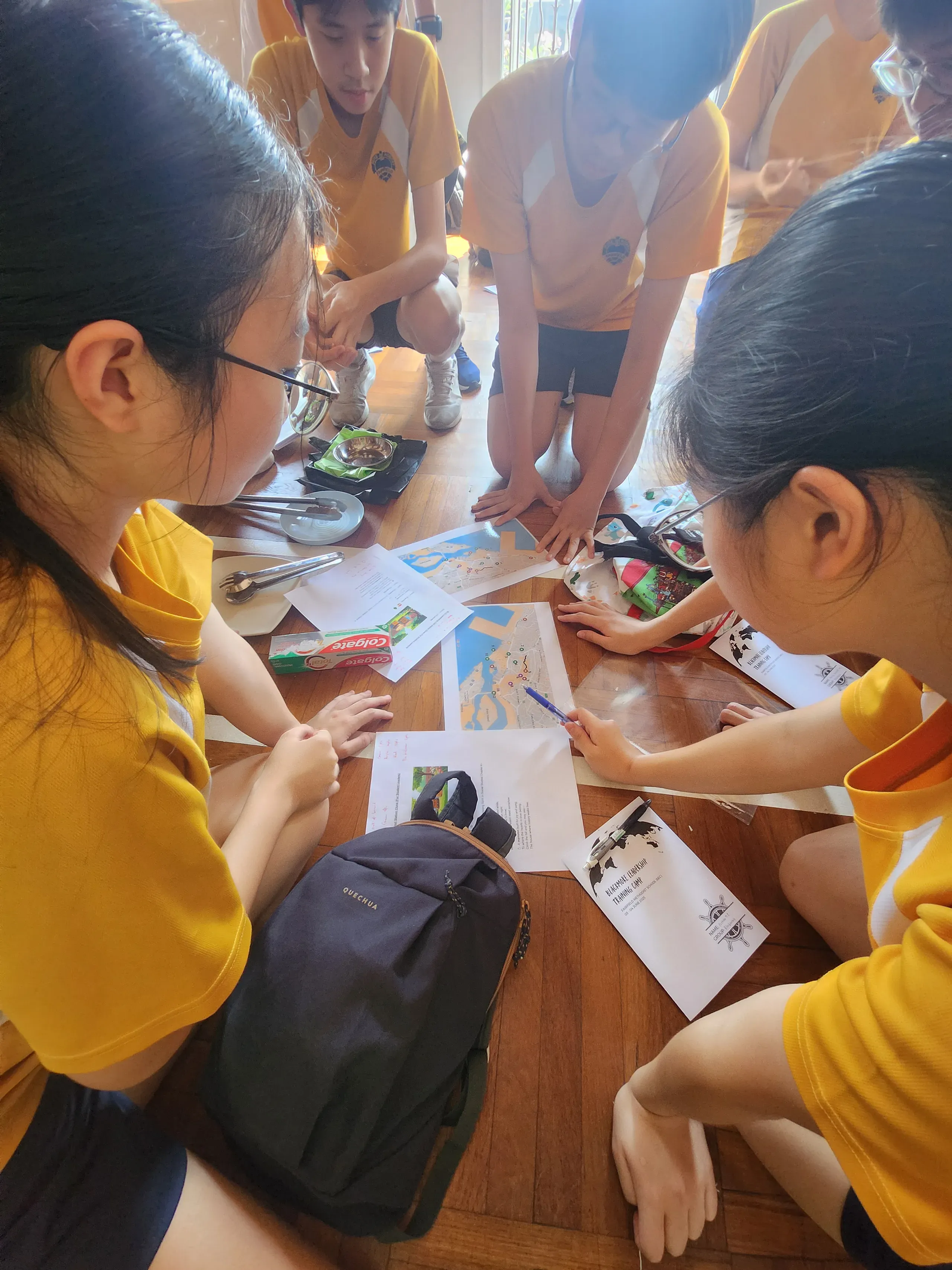 A high-angle close-up of five student leaders huddled in a tight circle on a wooden parquet floor, intensely focusing on a planning activity. Dressed in yellow and white school sports uniforms, they are reviewing printed maps, worksheets, and activity booklets spread across the floor. One student points a blue pen at a map while the others lean in closely to collaborate. A black backpack and scattered personal items sit on the floor beside them.