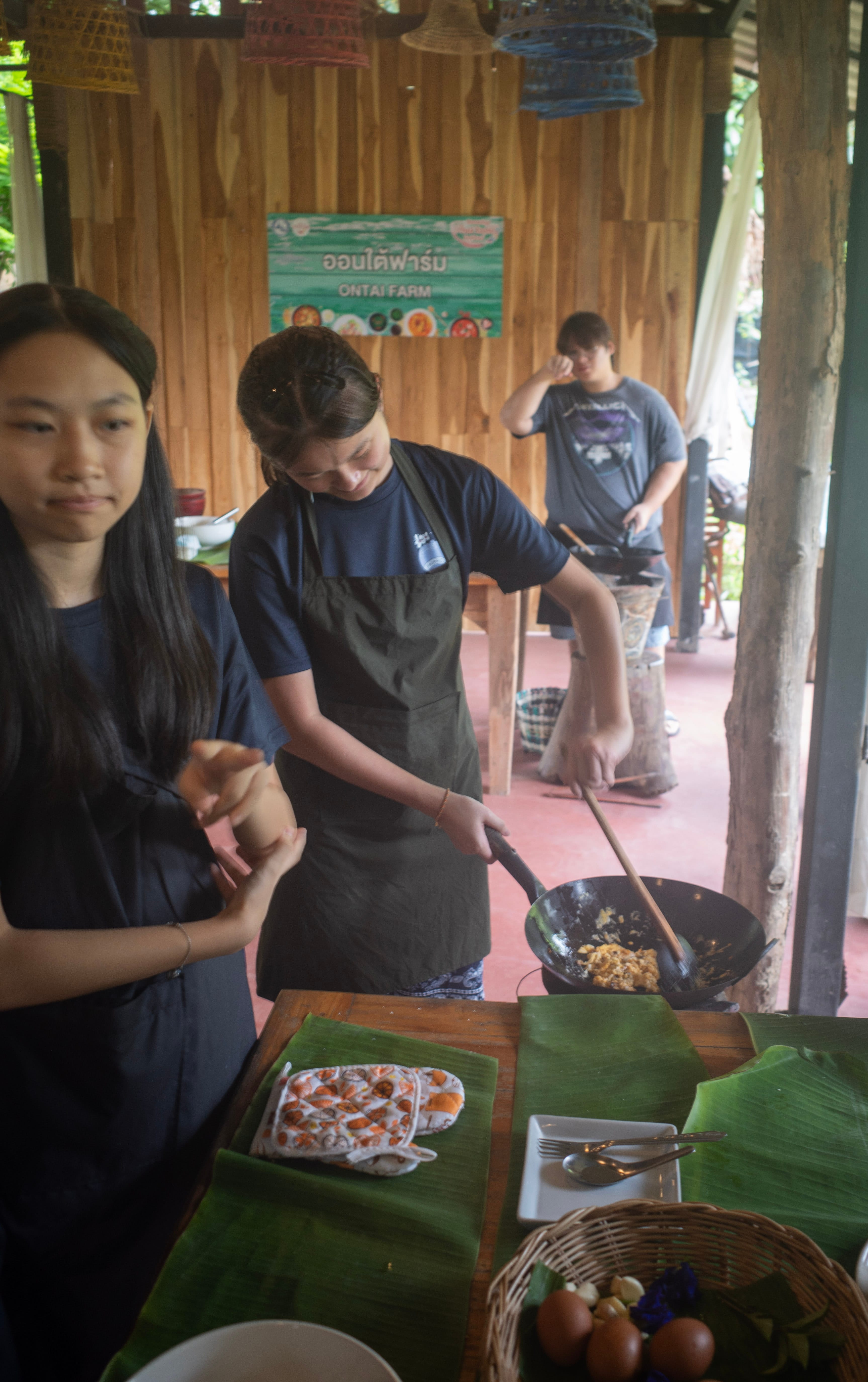Two female Secondary 3 students participate in a culinary class at an open-air pavilion kitchen at Ontai Farm. In the center, a student wearing a dark green apron looks down with a smile while using a wooden spatula to stir-fry ingredients in a large black wok on a portable stove. To her left, another student stands by observing. The foreground features a wooden table neatly set with large green banana leaves used as placemats, an oven mitt, a small white plate with cutlery, and a woven basket holding fresh eggs. In the background, a sign reads "ONTAI FARM" below Thai script on a wooden wall, with another student cooking at a separate station.