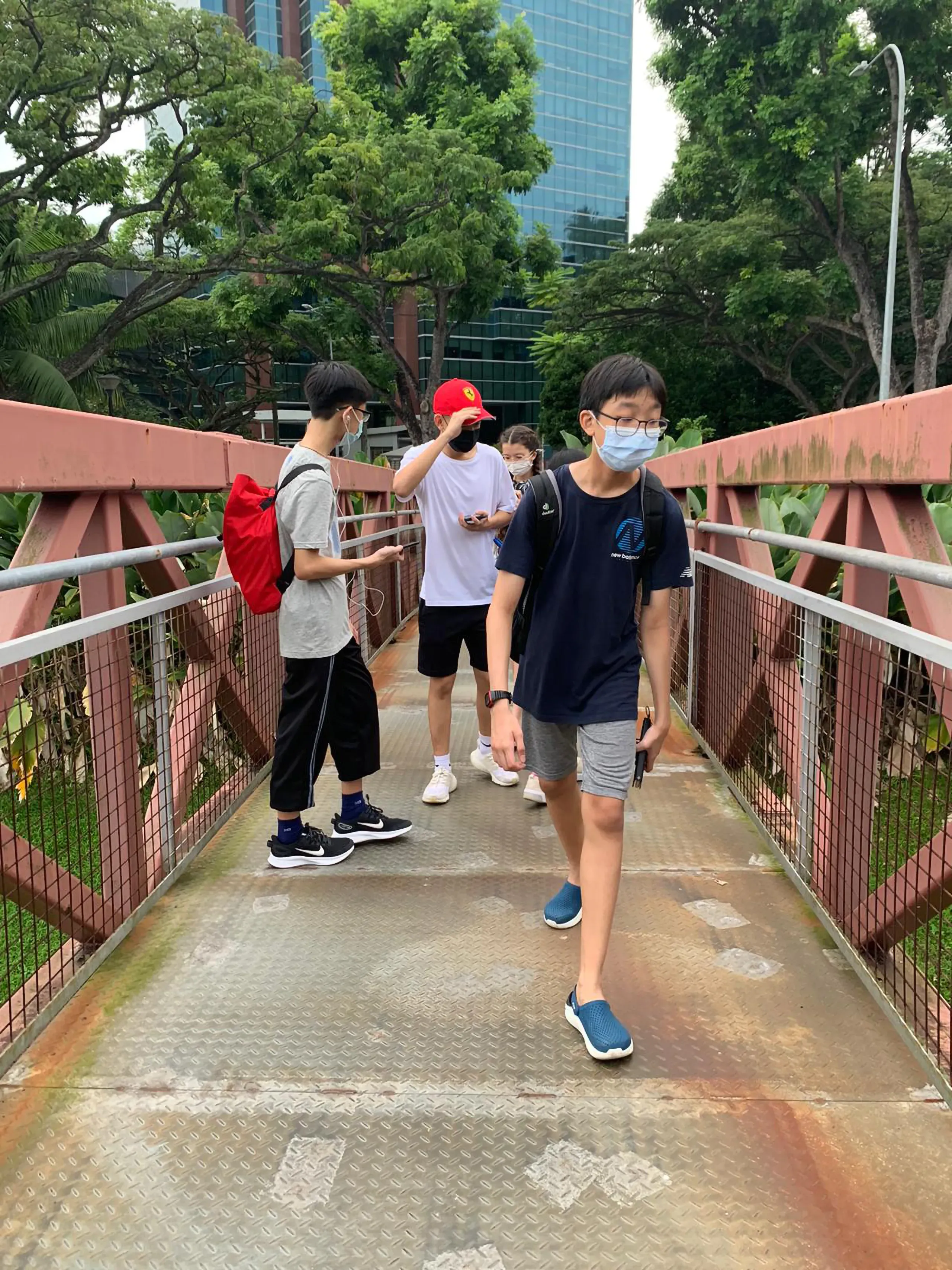 A group of Fairfield Methodist School (Secondary) students walking across an outdoor metal truss bridge near the campus during a school run activity. The students are dressed in casual t-shirts and shorts, wearing face masks and backpacks, with tall city buildings and lush green trees in the background.