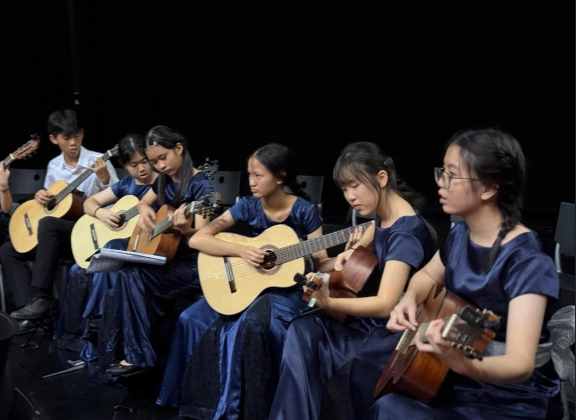 A close-up, side-angle view of a row of female students in the guitar ensemble performing on a dimly lit stage during SYF. The musicians are dressed in elegant, matching dark navy blue satin gowns and have their hair neatly braided. They are seated close together, intently focusing on playing their classical guitars. In the background on the far left, a male student in a white uniform shirt can also be seen playing his guitar.