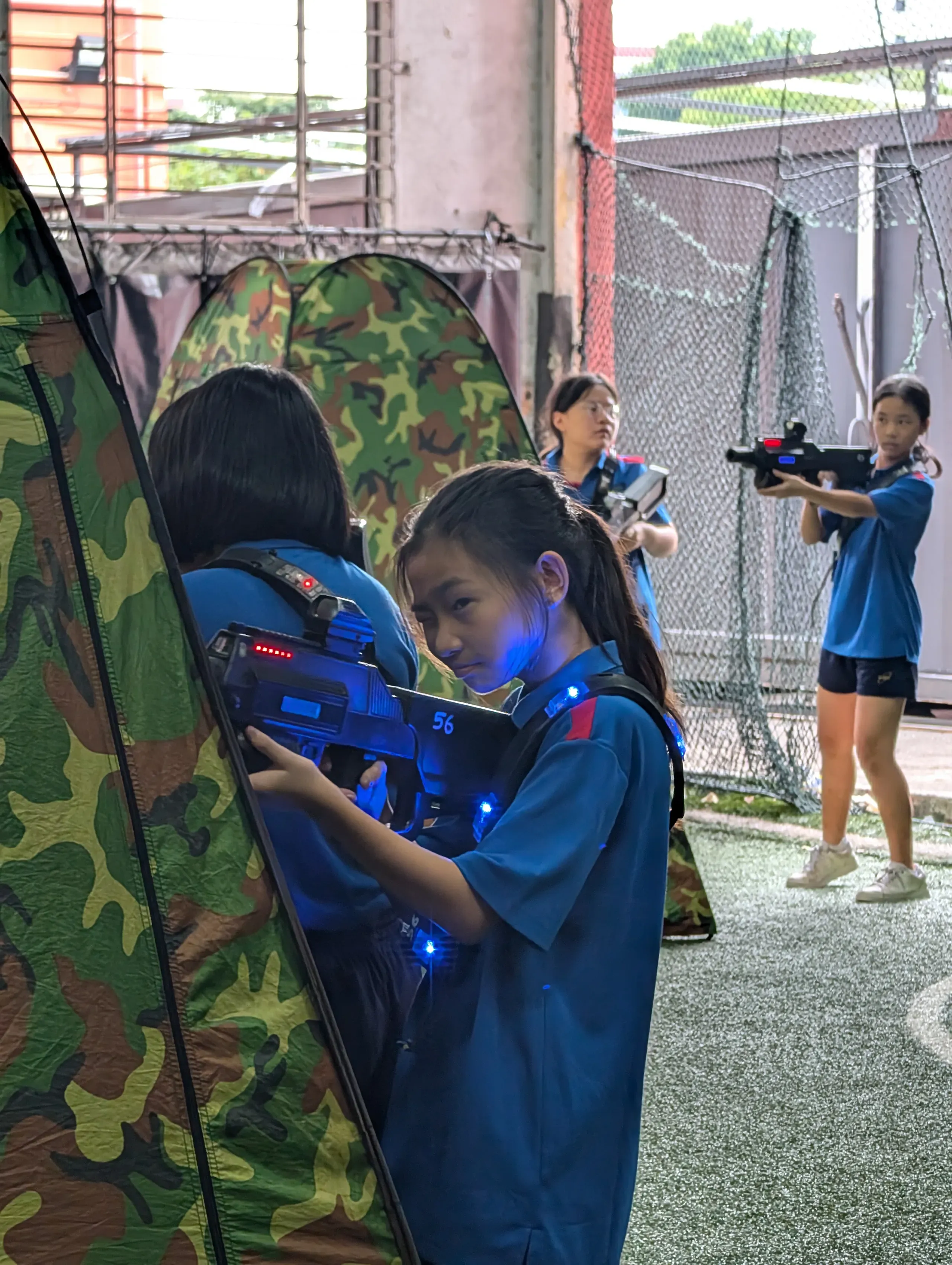 Four female Fairfield students in blue polo uniforms participate in a laser tag game on an indoor turf field. In the foreground, a girl with her hair in a braid takes cover behind a camouflage pop-up tent, holding a glowing blue laser tag blaster, while her teammates aim their blasters in the background.