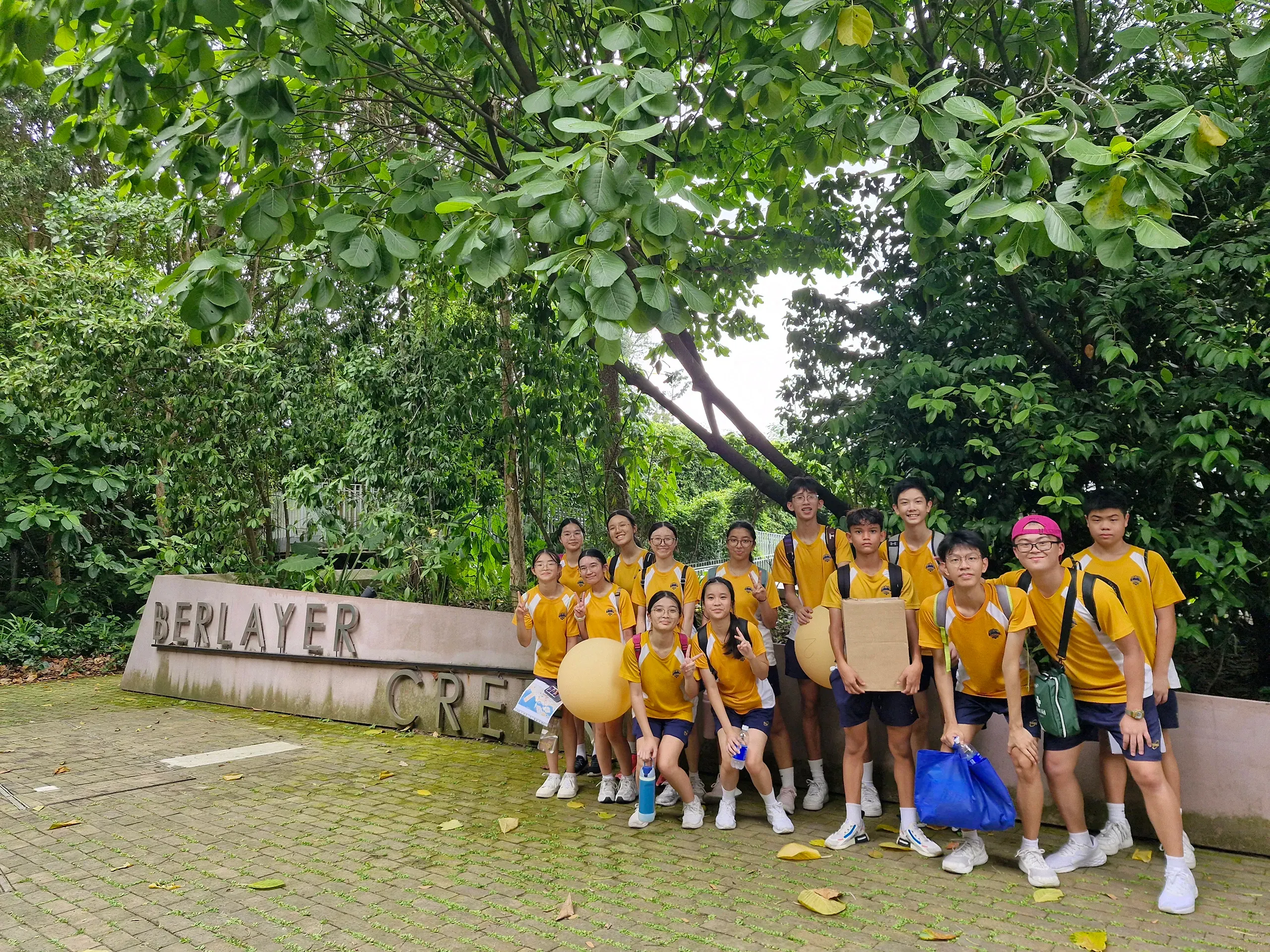 A group of fourteen student leaders posing together on a brick-paved path next to a low concrete sign that reads "BERLAYER CREEK" during their leadership camp. The students are dressed in yellow and white school sports t-shirts and dark shorts, with some carrying large cardboard boxes, a blue bag, and giant pale yellow balloons. They are all smiling and gesturing with peace signs under a dense canopy of lush green trees, with a hint of a concrete overpass structure peaking through the foliage above.
