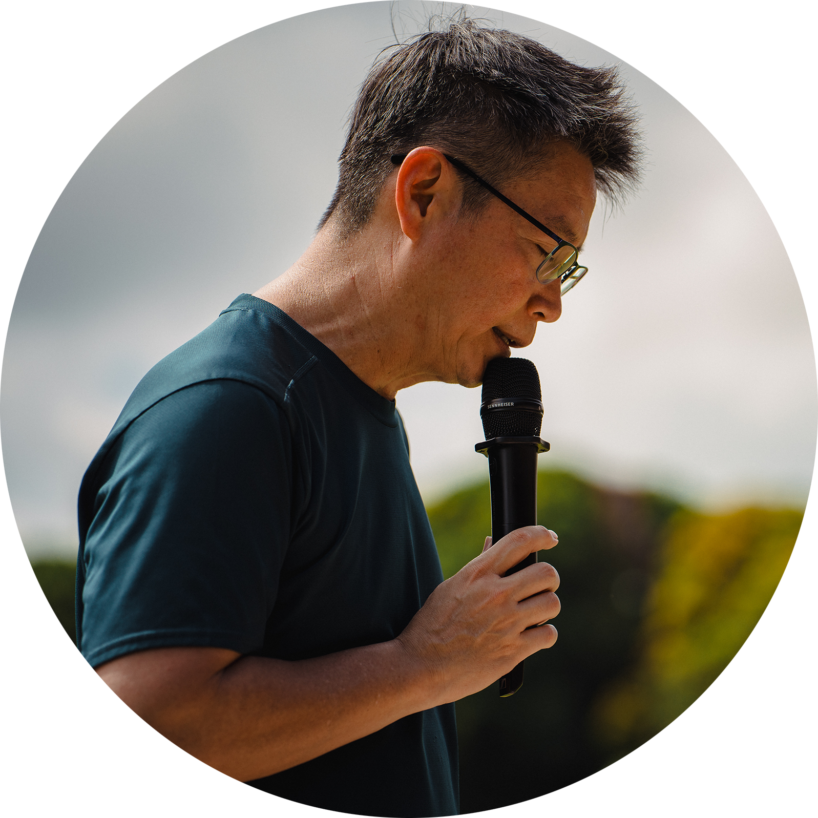 Side profile of the principal, Mr. Wee Tat Chuen, holding a microphone with eyes closed in prayer during school Sports Day, with a soft outdoor background.