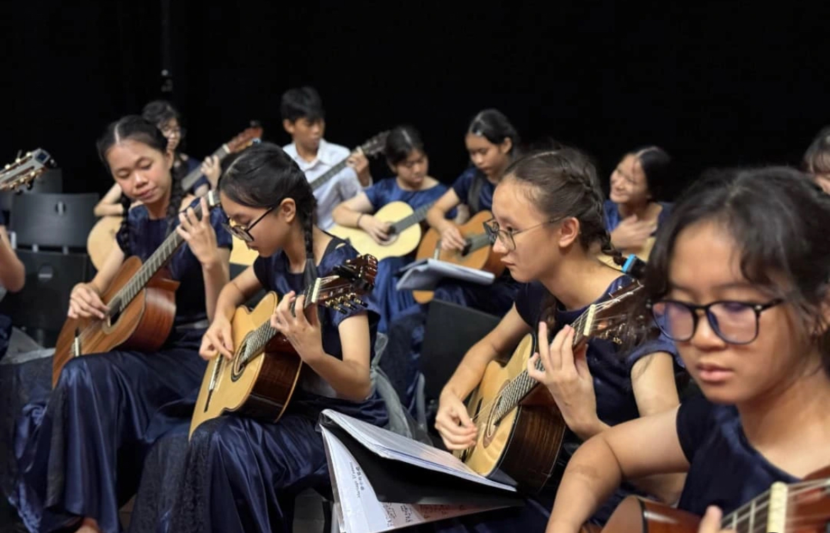 The school guitar ensemble performs on stage inside a grand, wood-paneled auditorium for the Singapore Youth Festival (SYF). The student musicians are seated in neat, semi-circular rows on a polished wooden stage, dressed in light yellow school shirts, ties, and blue skirts or dark trousers. They are all playing classical guitars resting on their left legs, reading from sheet music placed on black music stands in front of them. In the center foreground, a conductor stands on an elevated wooden podium with their back to the empty, red-upholstered theater seats, directing the performance under bright overhead stage lights.