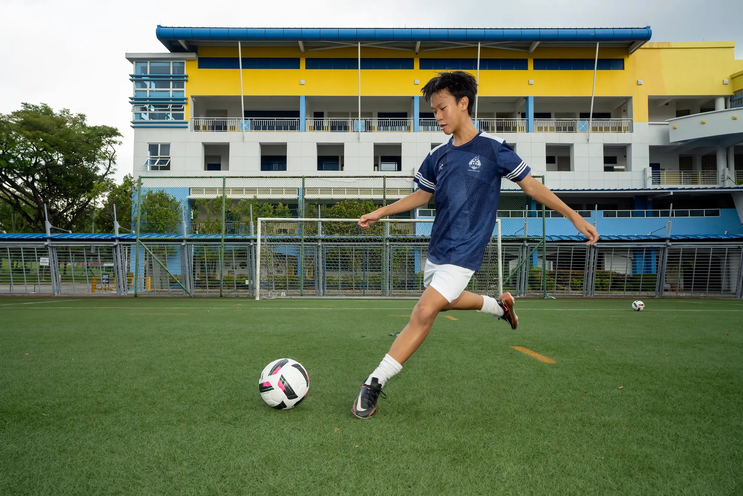 A male student in a dark blue patterned soccer jersey and white shorts kicking a football on a green artificial turf field, with a prominent yellow and blue school building and a goalpost in the background.