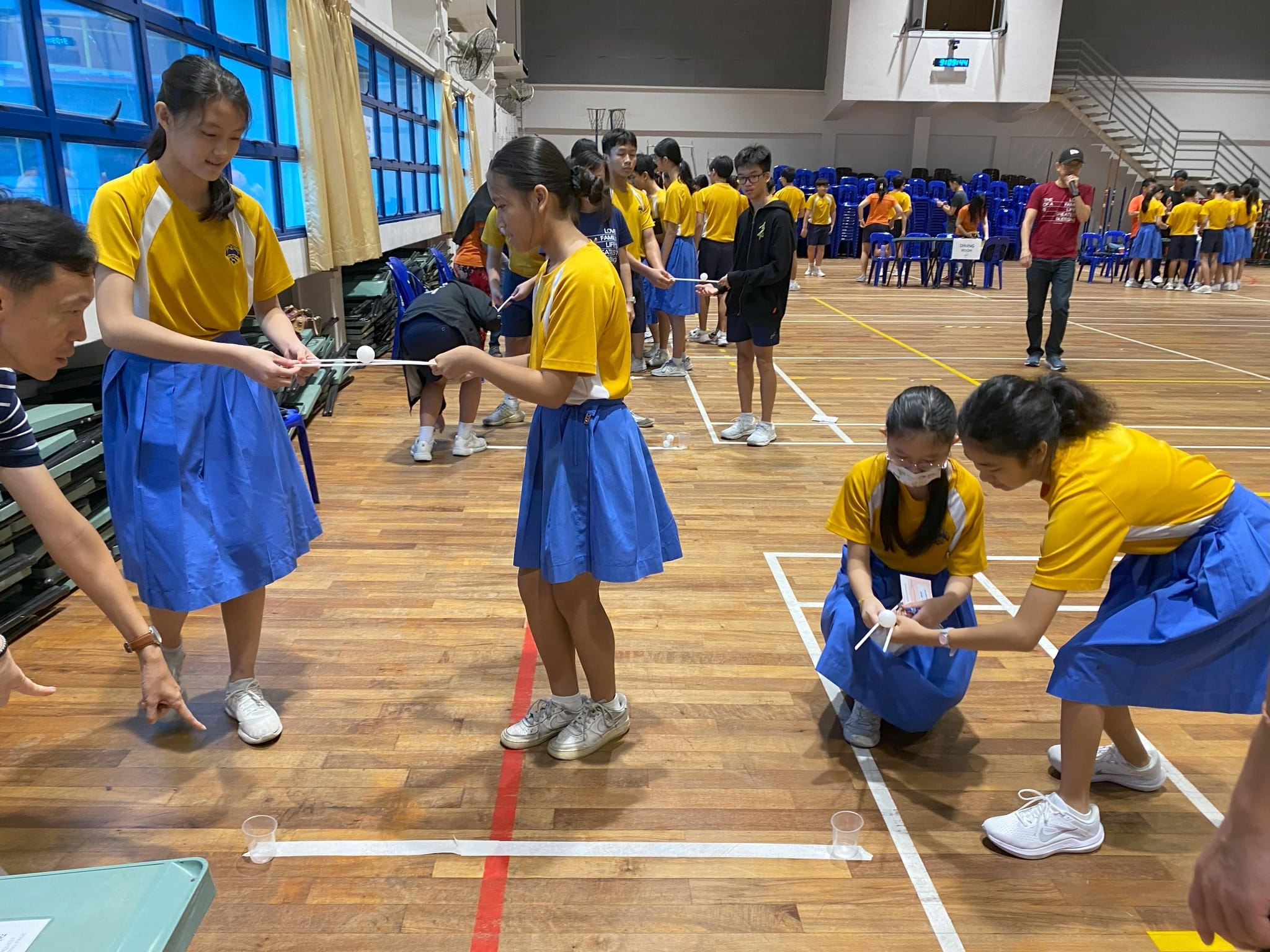 Secondary Two students participate in a coordinated game during the FamQuest program in the school hall for FLEX week. Wearing their yellow-and-white school PE shirts and blue skirts, two female students on the left stand facing each other, using long white sticks to steady a ping-pong ball. On the right, another pair of female students kneel down to carefully balance a ping-pong ball using chopsticks. On the far left, a parent facilitator leans forward, pointing toward the floor lines to guide the students through the challenge.